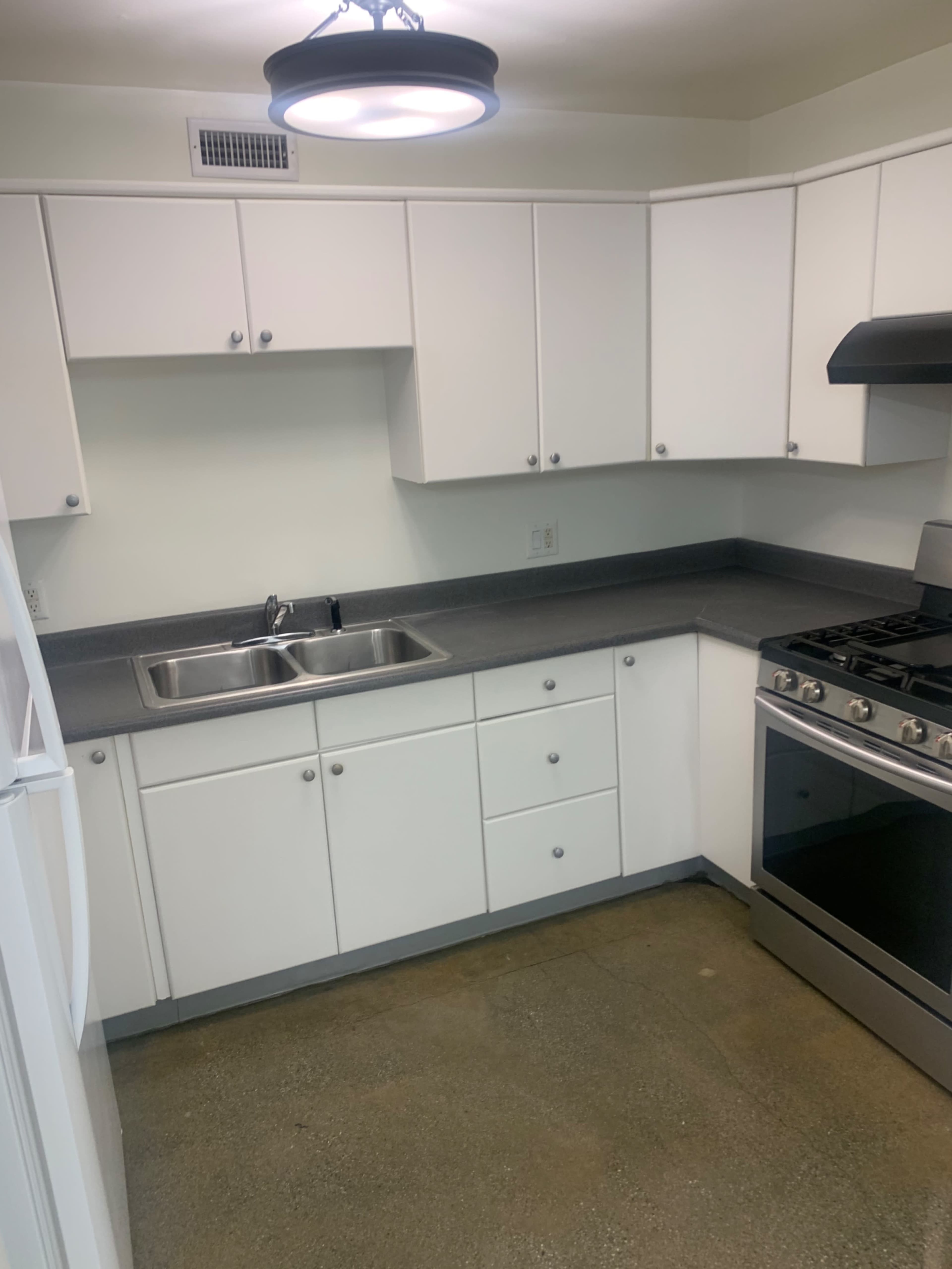 The image shows a modern kitchen with white cabinets, a stainless steel stove, and a double sink, featuring gray countertops and a concrete floor.