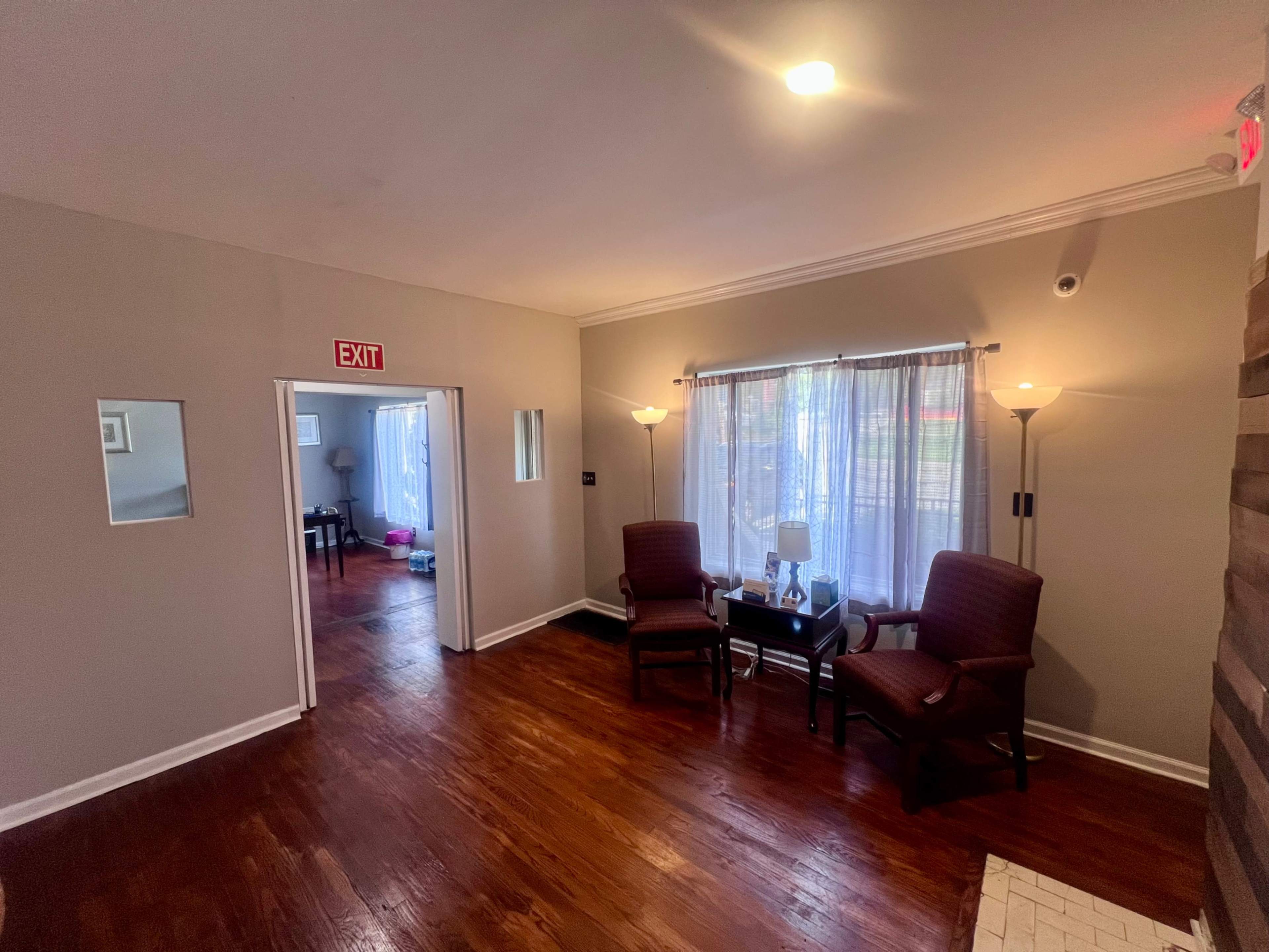 The image shows a cozy waiting area with two chairs, a small table, and a bright window illuminating the space.