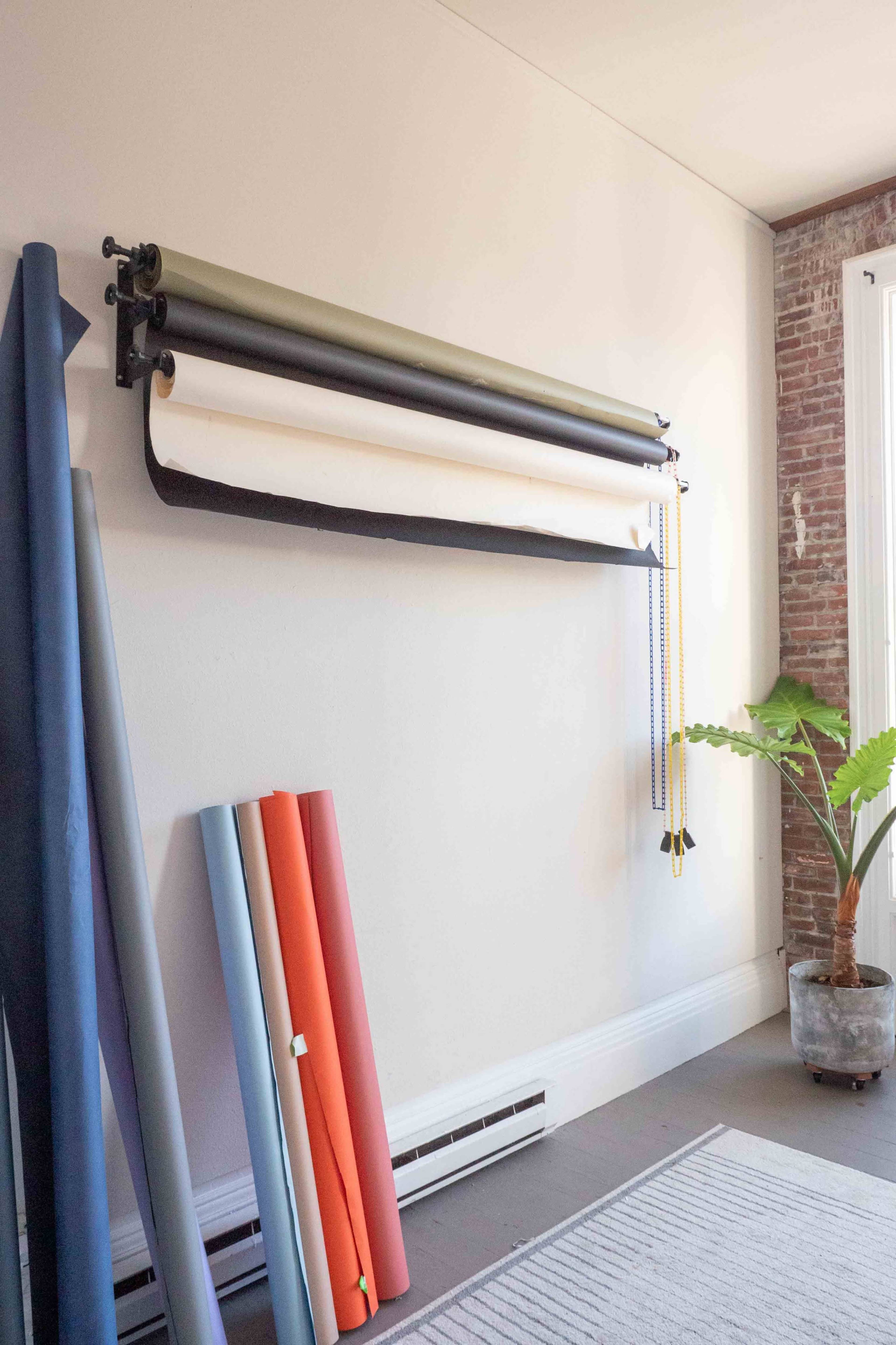A wall-mounted roll of photography backdrop paper is flanked by neatly stacked rolls of colored paper and a green potted plant in a spacious room.