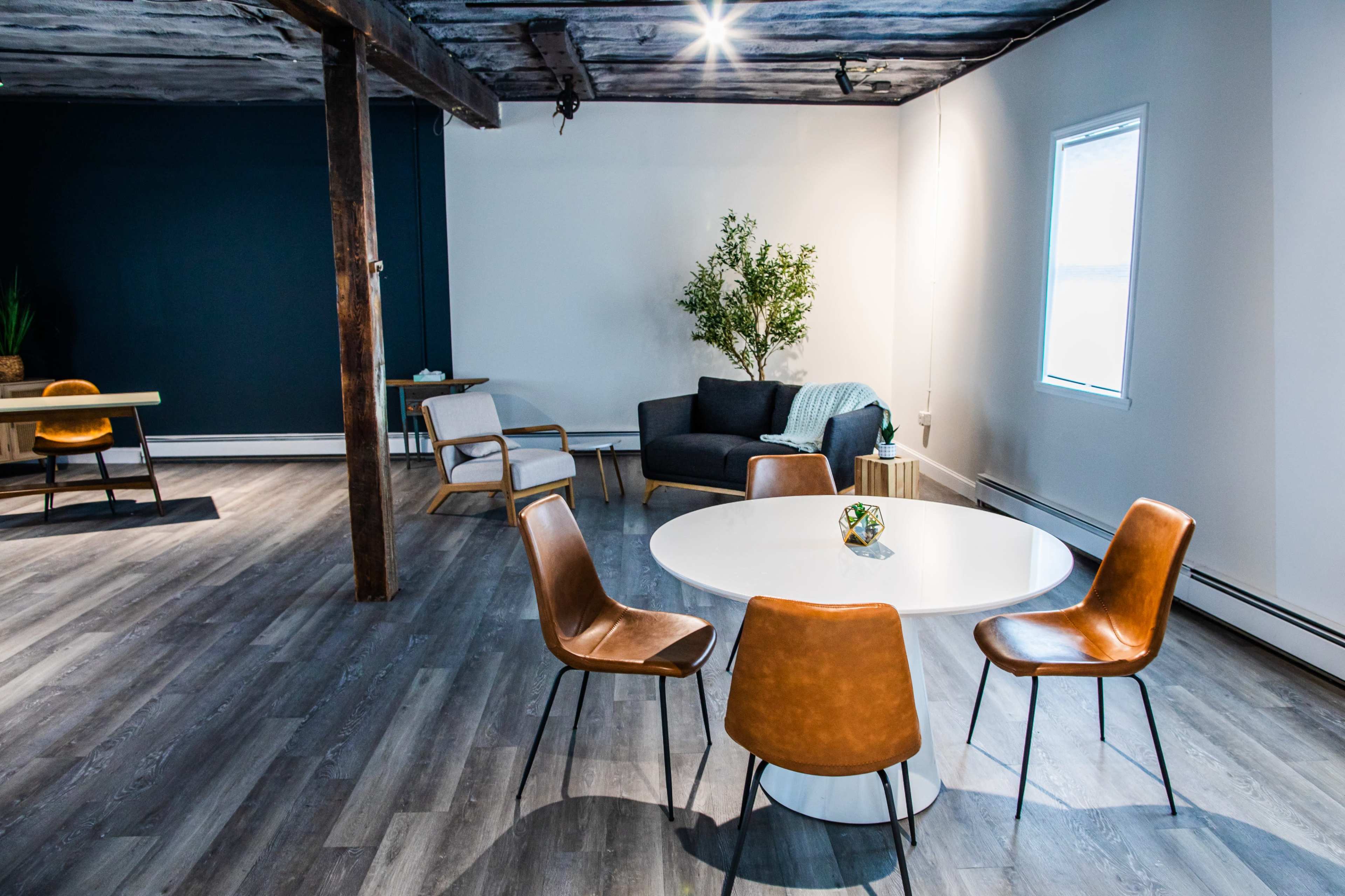 The image shows a modern, minimally decorated room featuring a round white table with four brown chairs, a gray couch, and a potted plant.