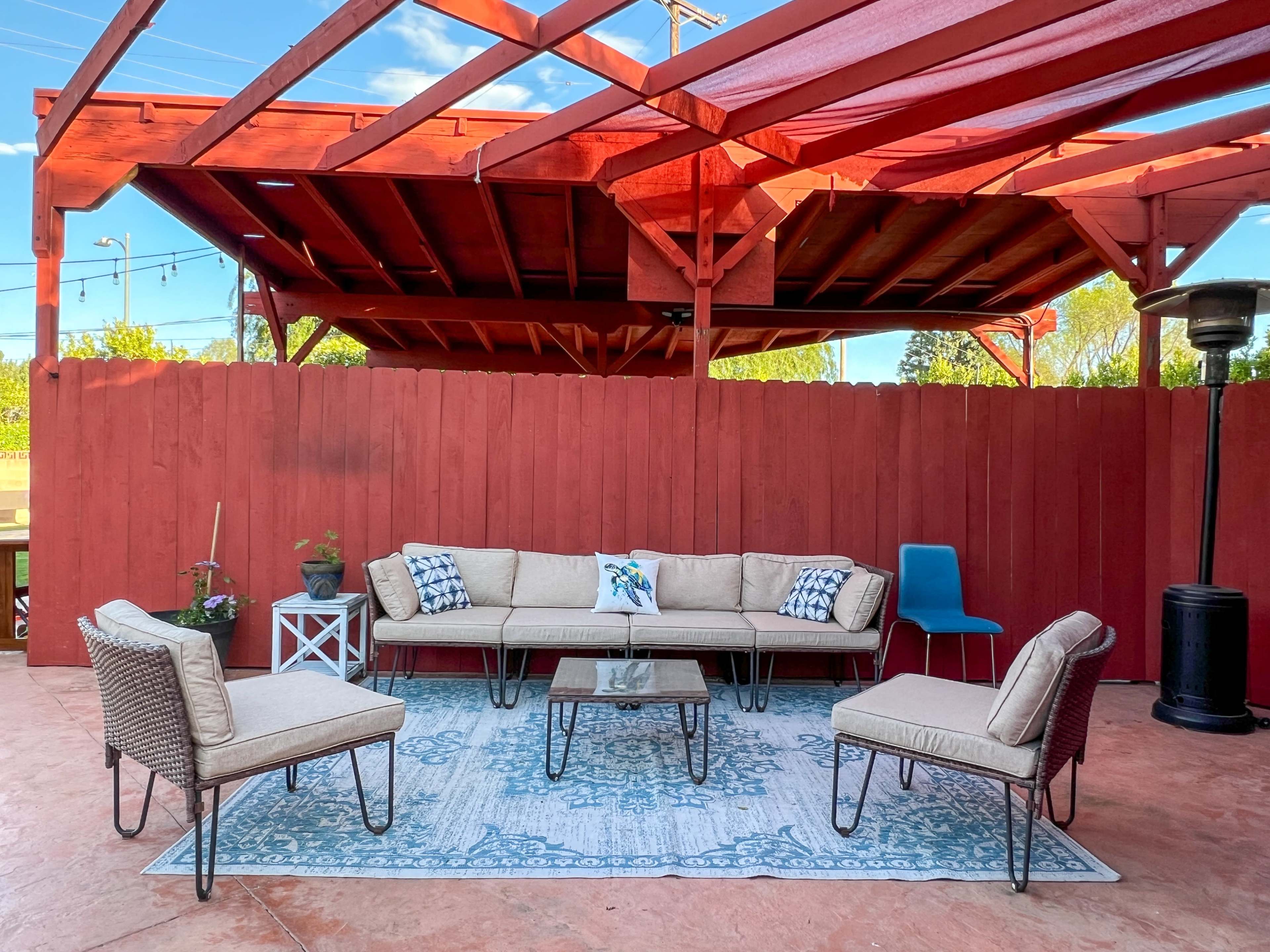 A patio seating area features a beige couch, chairs, and a table arranged on a decorative rug beneath a wooden pergola with a red fence in the background.