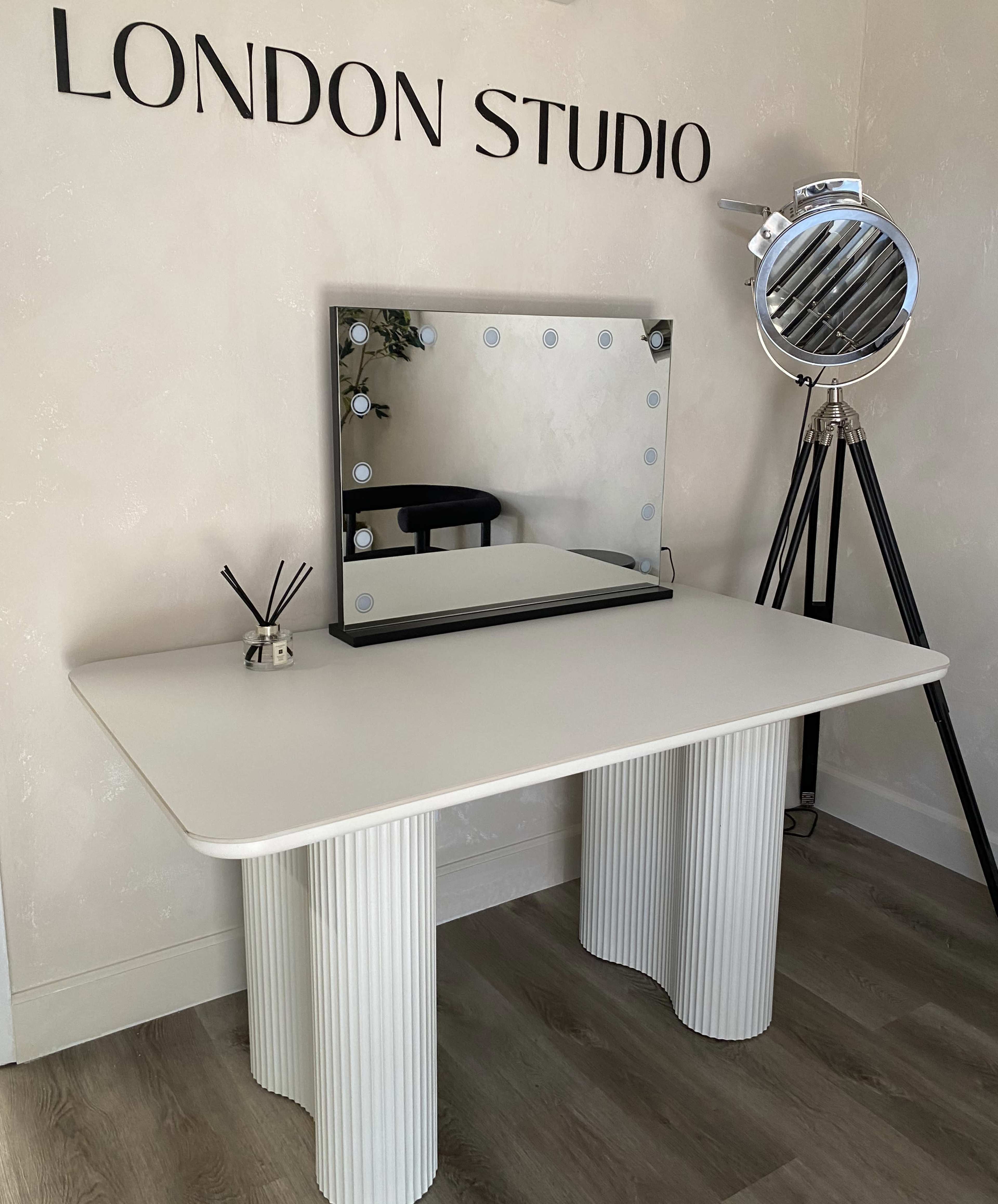 A modern white table with fluted columns, a round mirror with lights, and a vintage-style spotlight in a room labelled "LONDON STUDIO."