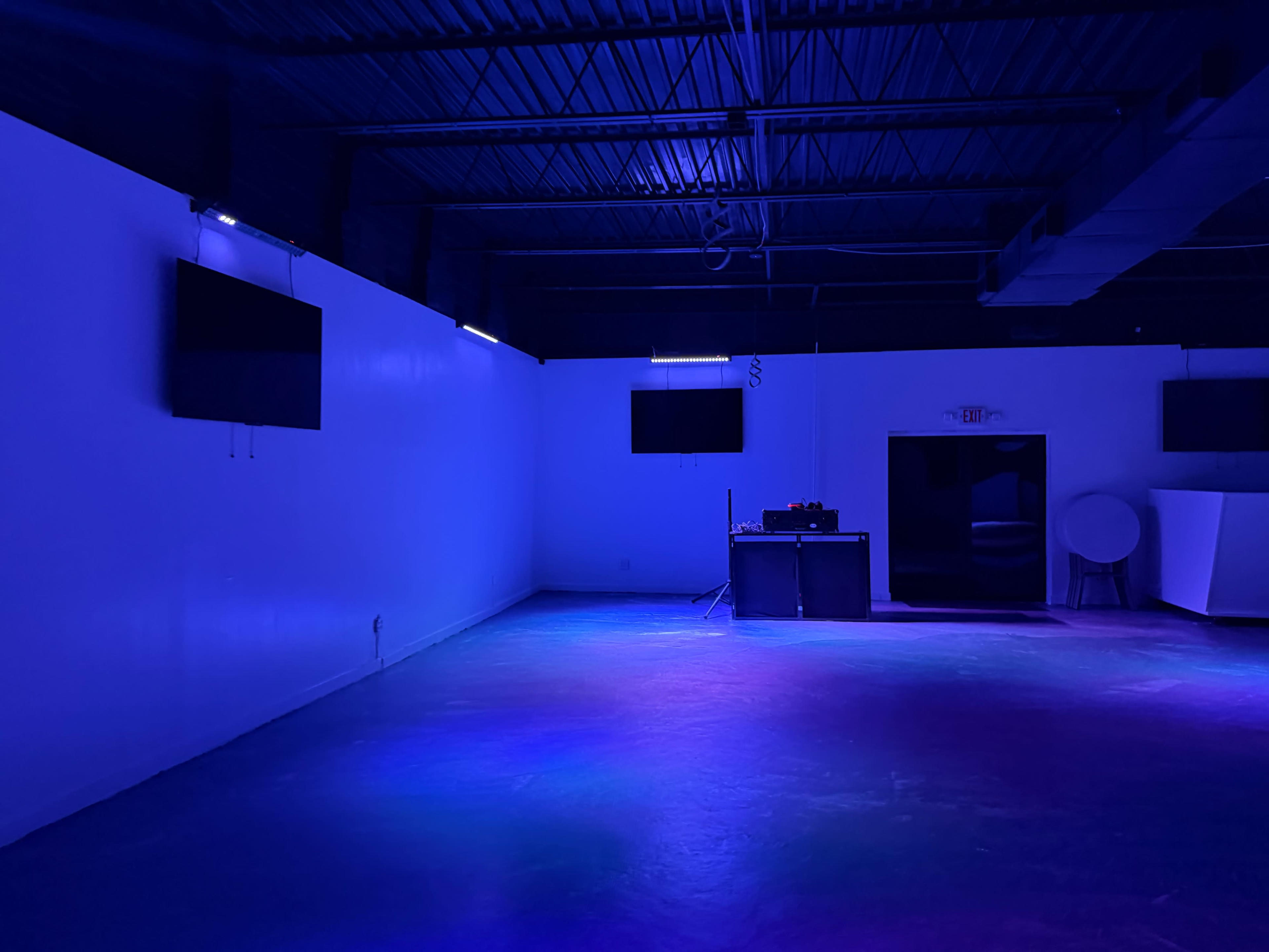 The image shows an empty room with blue lighting, featuring two wall-mounted screens, a small bar area, and a black door leading out.