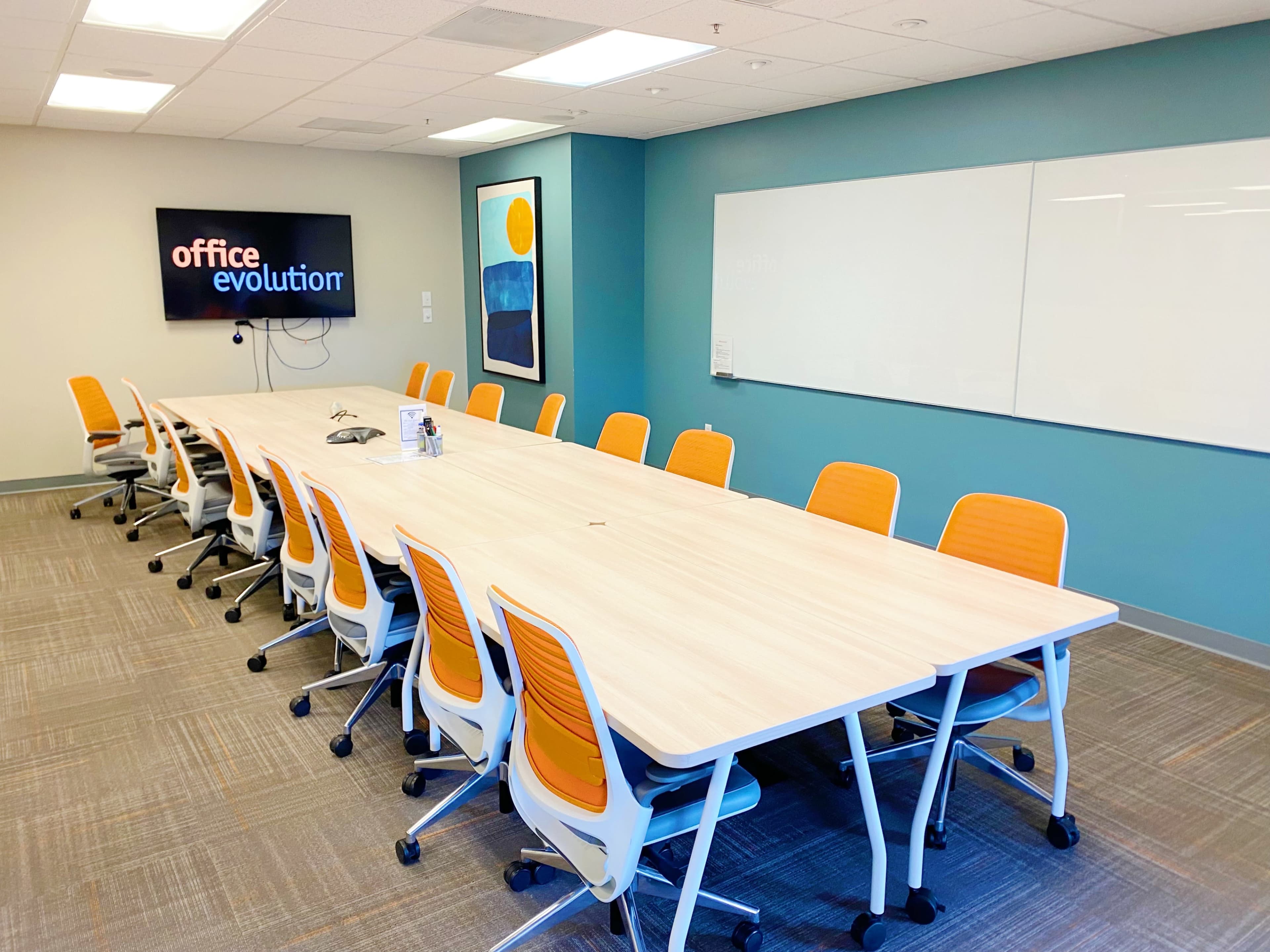 A large conference room features a long wooden table surrounded by orange and white chairs, with a screen displaying "office evolution" on the wall and a whiteboard nearby.