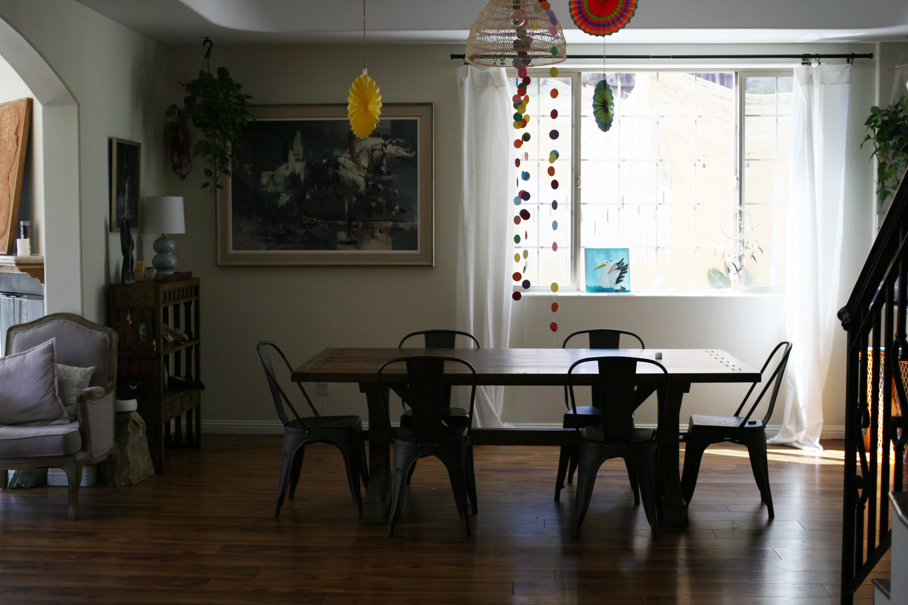 A dining area features a wooden table surrounded by black chairs, with decorative elements hanging from the ceiling and a large window letting in natural light.
