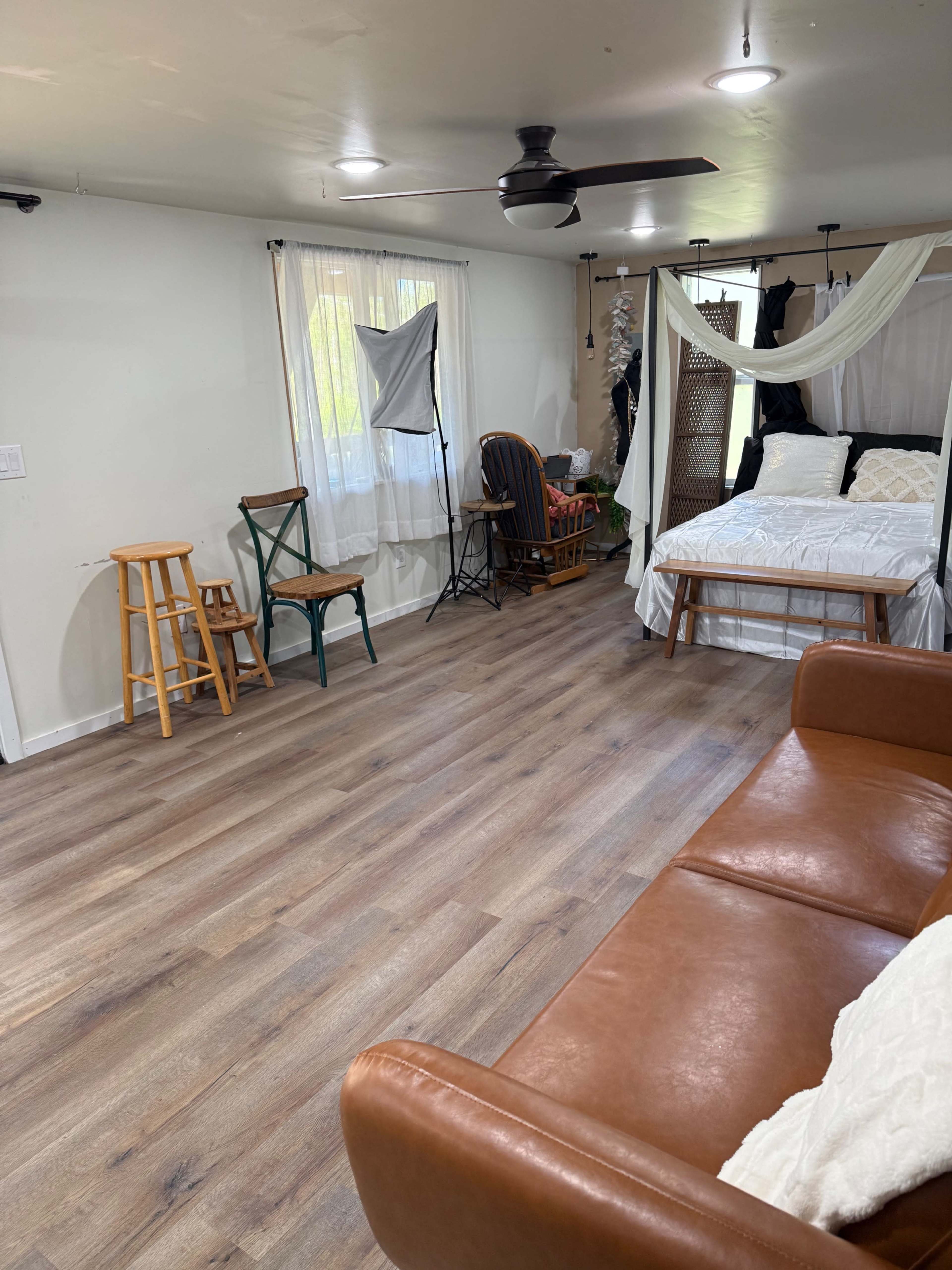 The image shows a spacious room with wooden flooring, featuring a bed with white bedding, a couch, various chairs, and photography equipment set up near the window.
