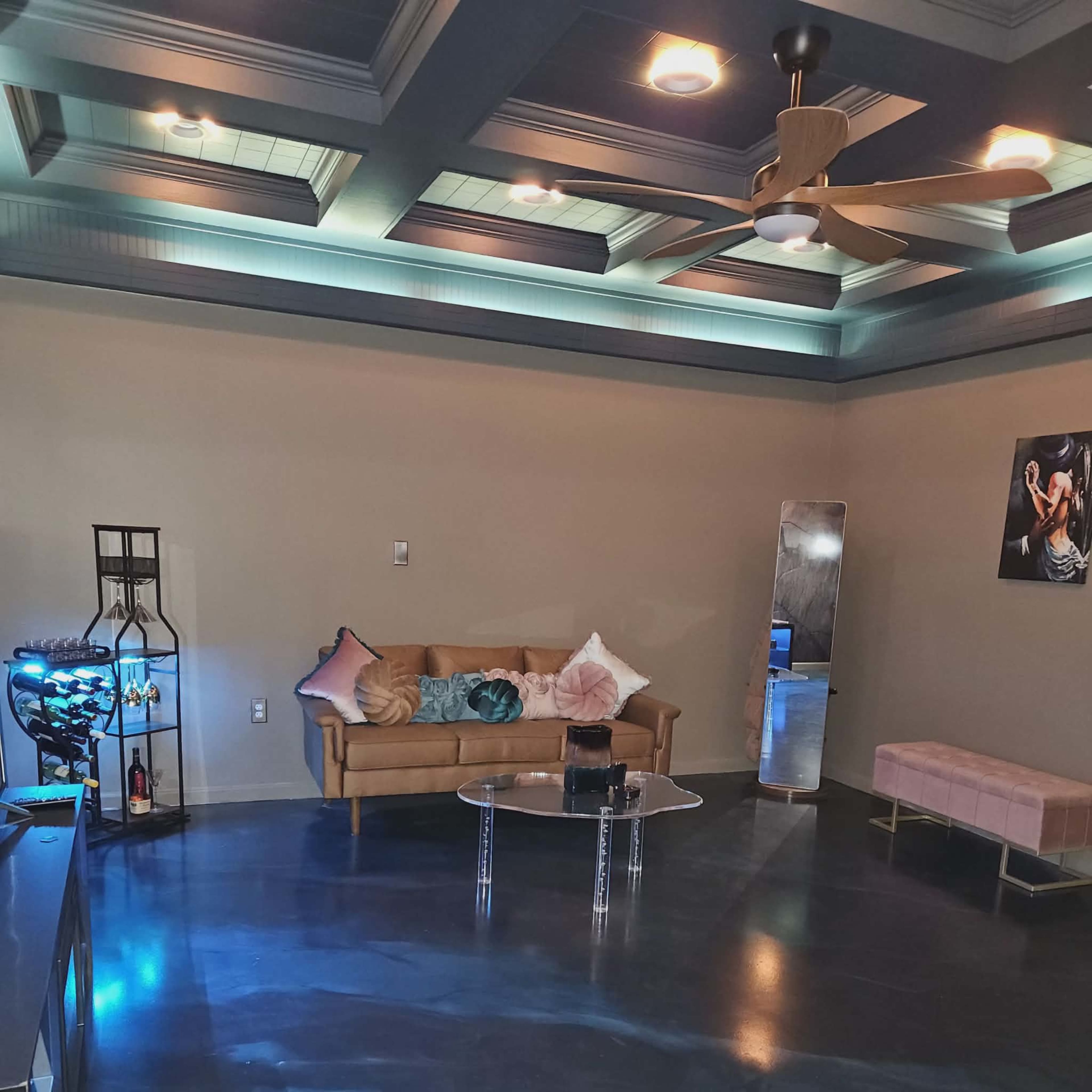 A spacious room features a tan sofa with decorative pillows, a glass coffee table, a bench, a ceiling fan, and modern lighting.