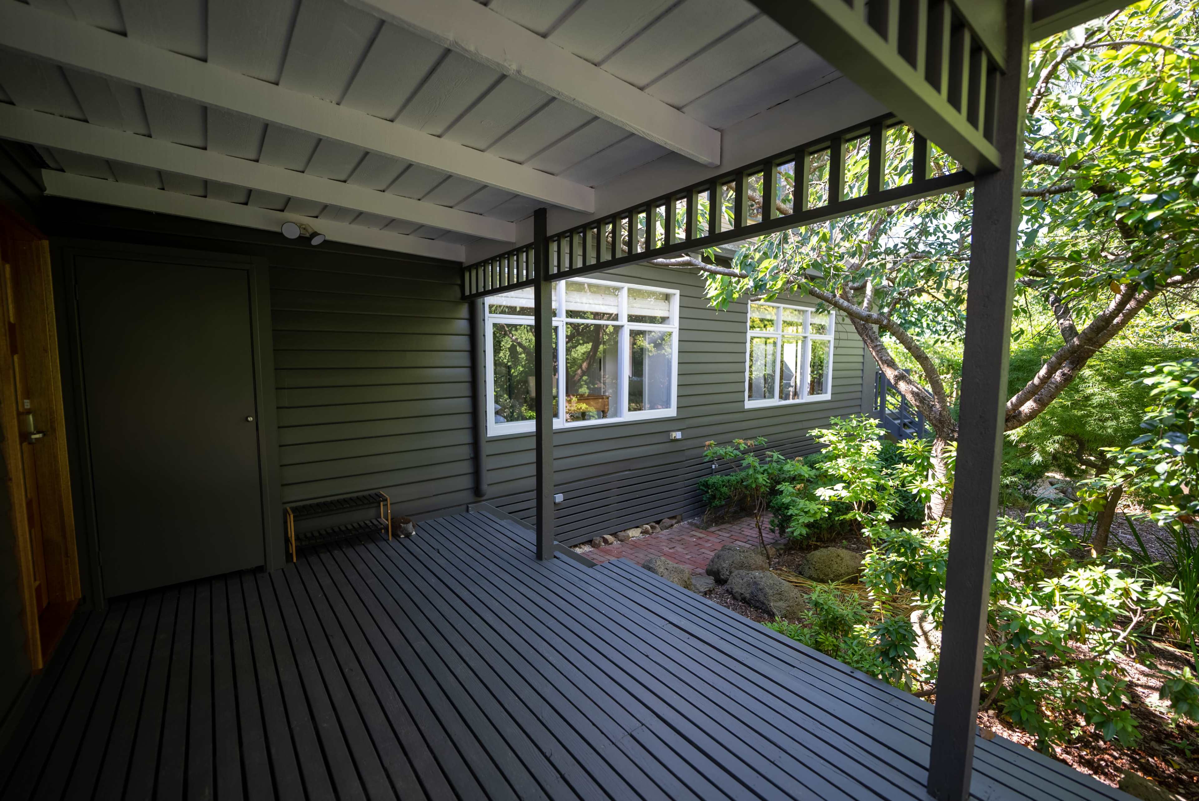 A shaded porch with wooden flooring leads to a house featuring green siding and large windows surrounded by lush foliage.