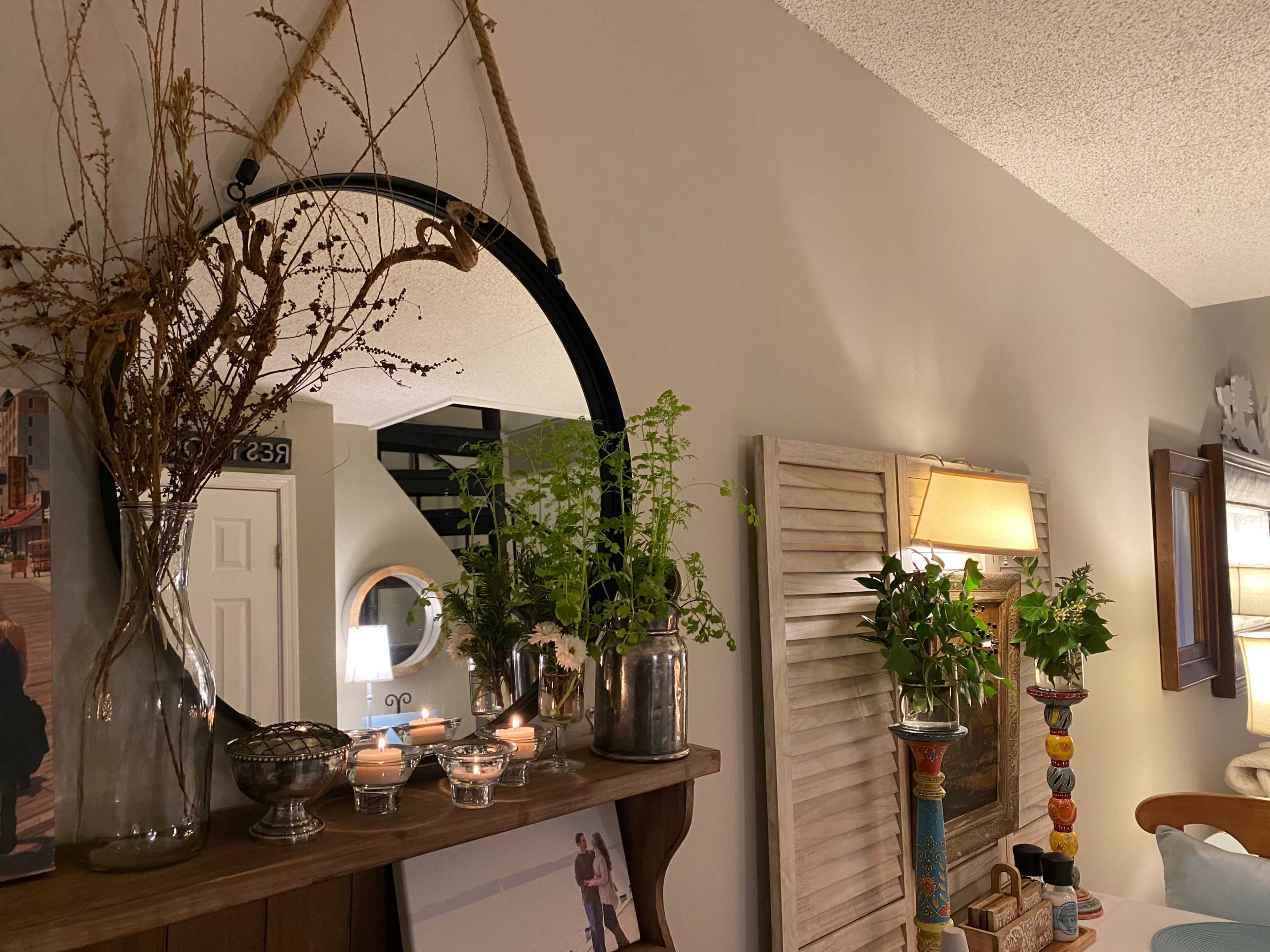 A circular mirror hangs on a wall adorned with plants and decorative items, while a framed photo sits on a wooden shelf below.