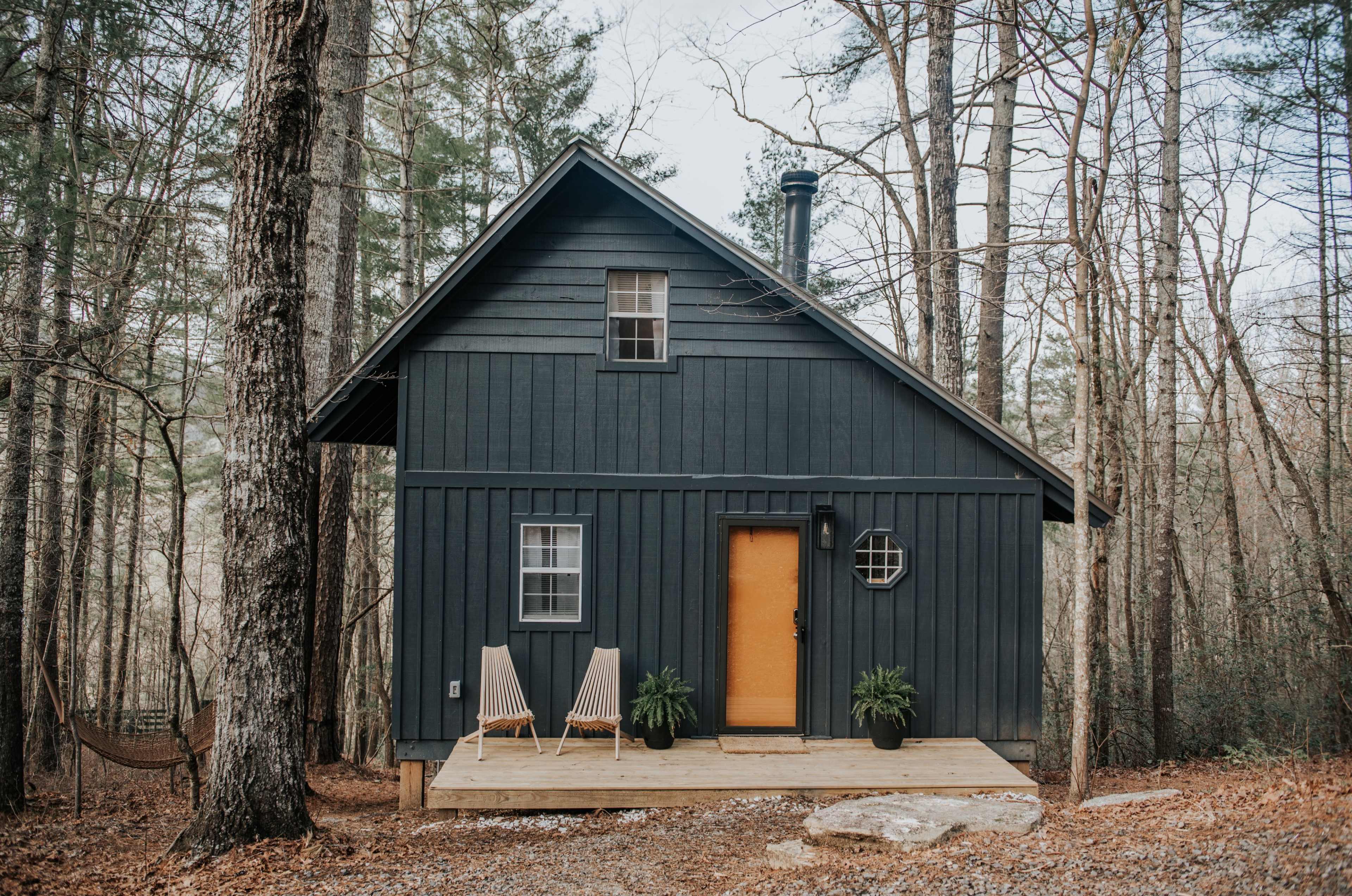 A dark wooden cabin with a yellow door and two chairs sits on a wooden porch surrounded by trees.