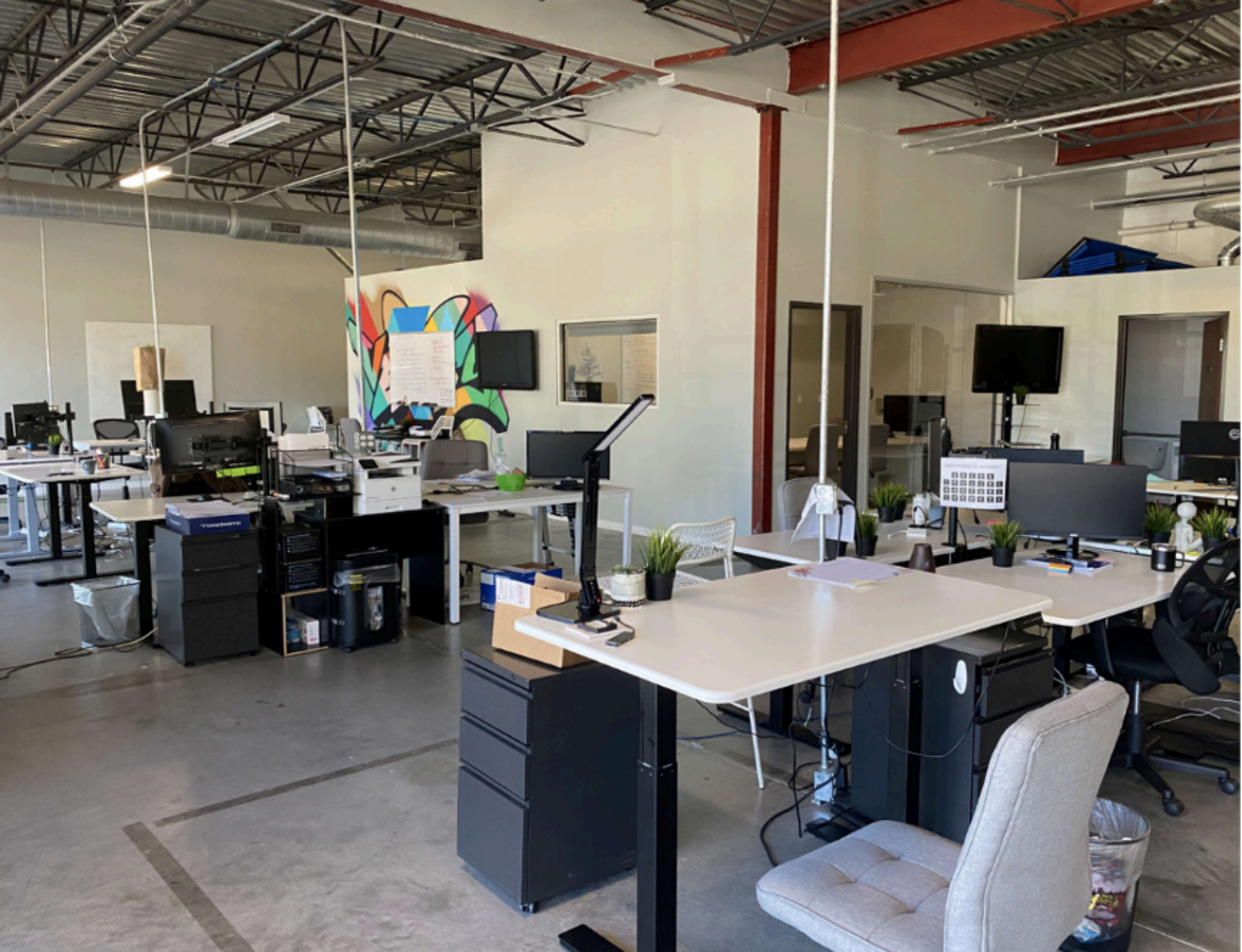 The image shows a spacious, modern office with several workstations featuring desks, chairs, and plants, along with a colorful mural on one wall.