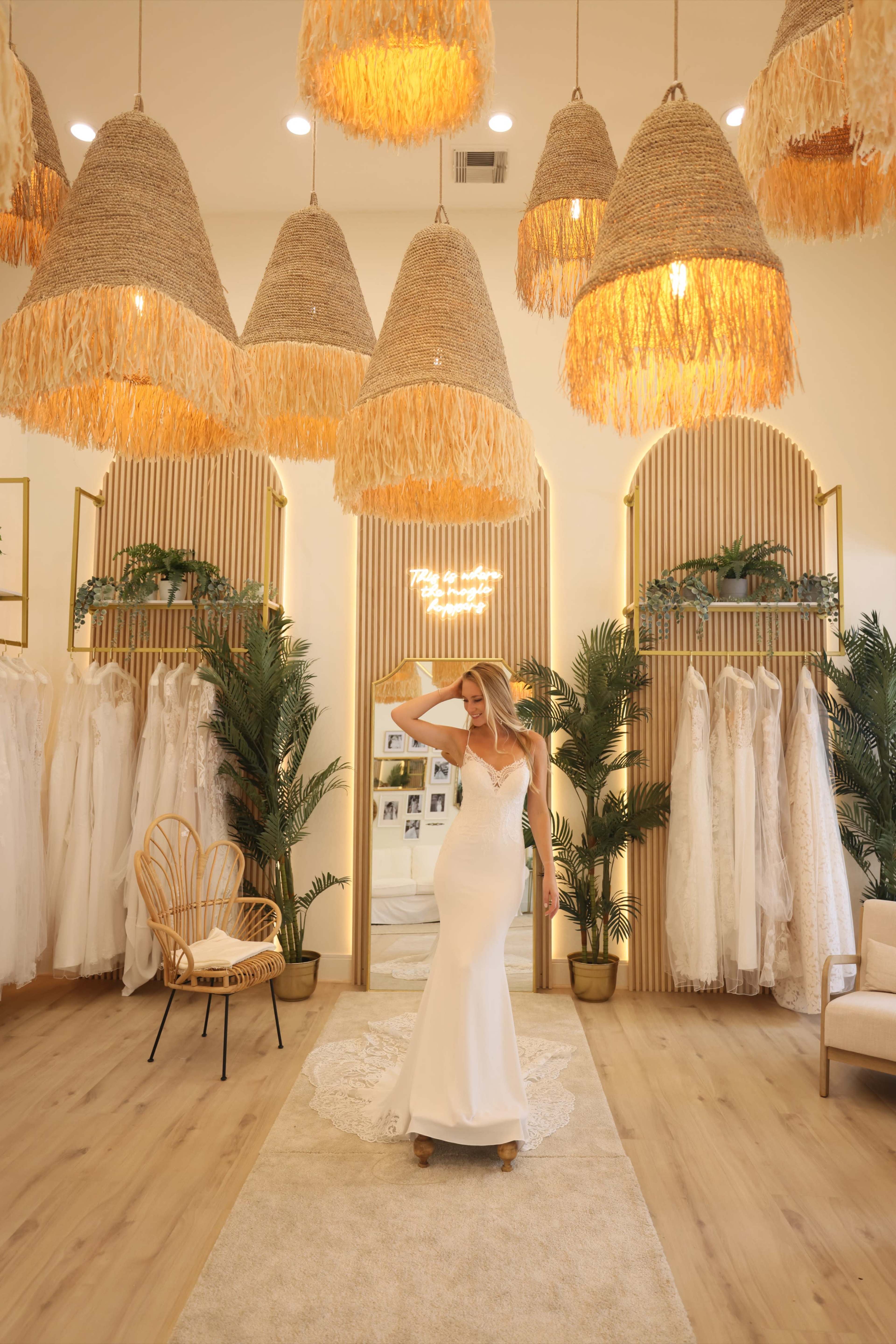 A bride poses in a bridal boutique surrounded by hanging light fixtures and racks of wedding dresses.