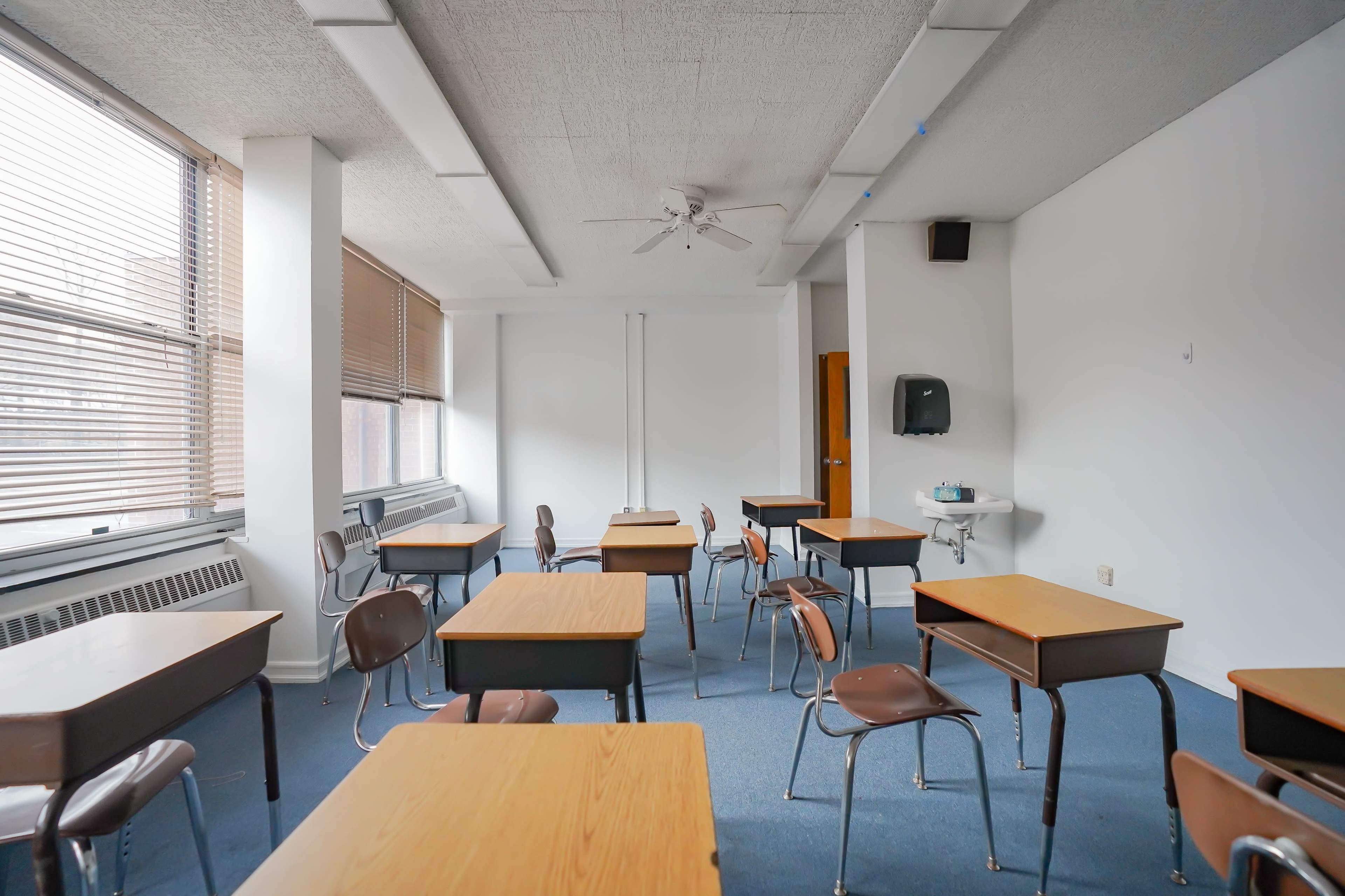A classroom with wooden desks and chairs arranged in an open layout, featuring large windows and a sink in the corner.