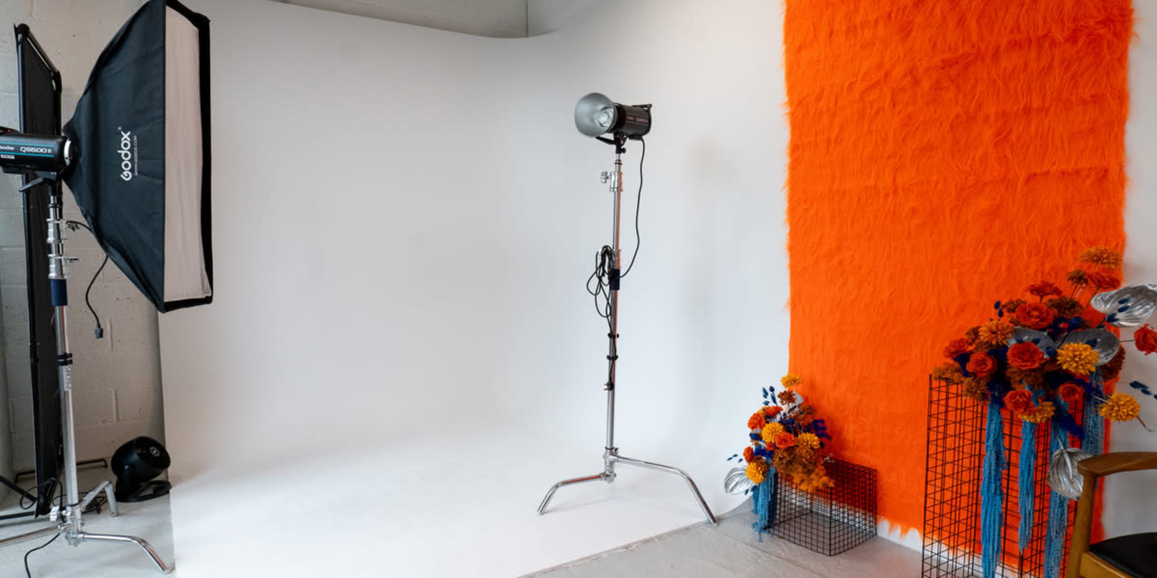 The image shows a photography studio featuring a large orange backdrop, a softbox light on a stand, and a decorative arrangement of flowers in a wire basket.