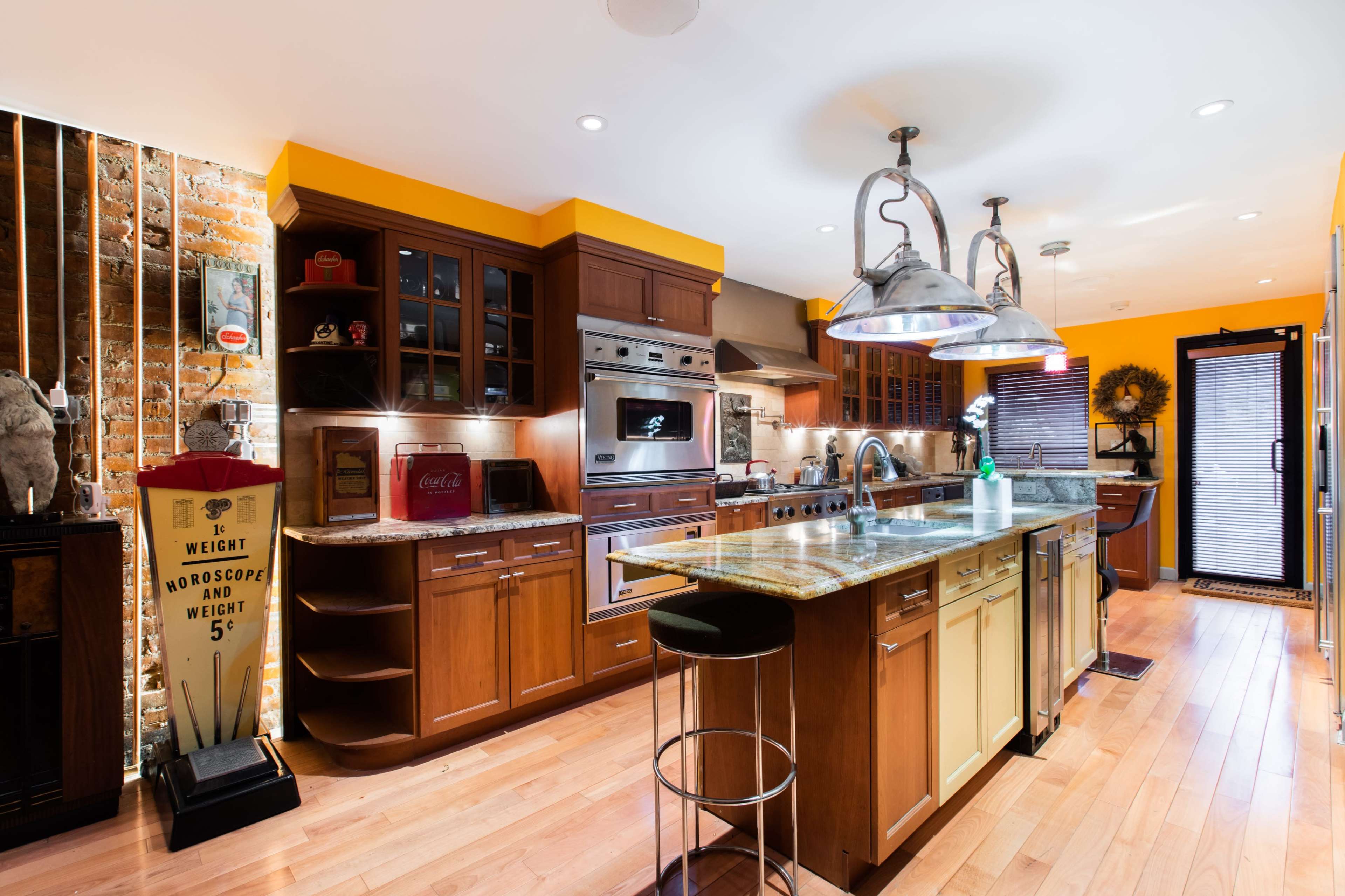 A modern kitchen featuring wooden cabinets, granite countertops, industrial-style light fixtures, and a mix of orange and brick walls.