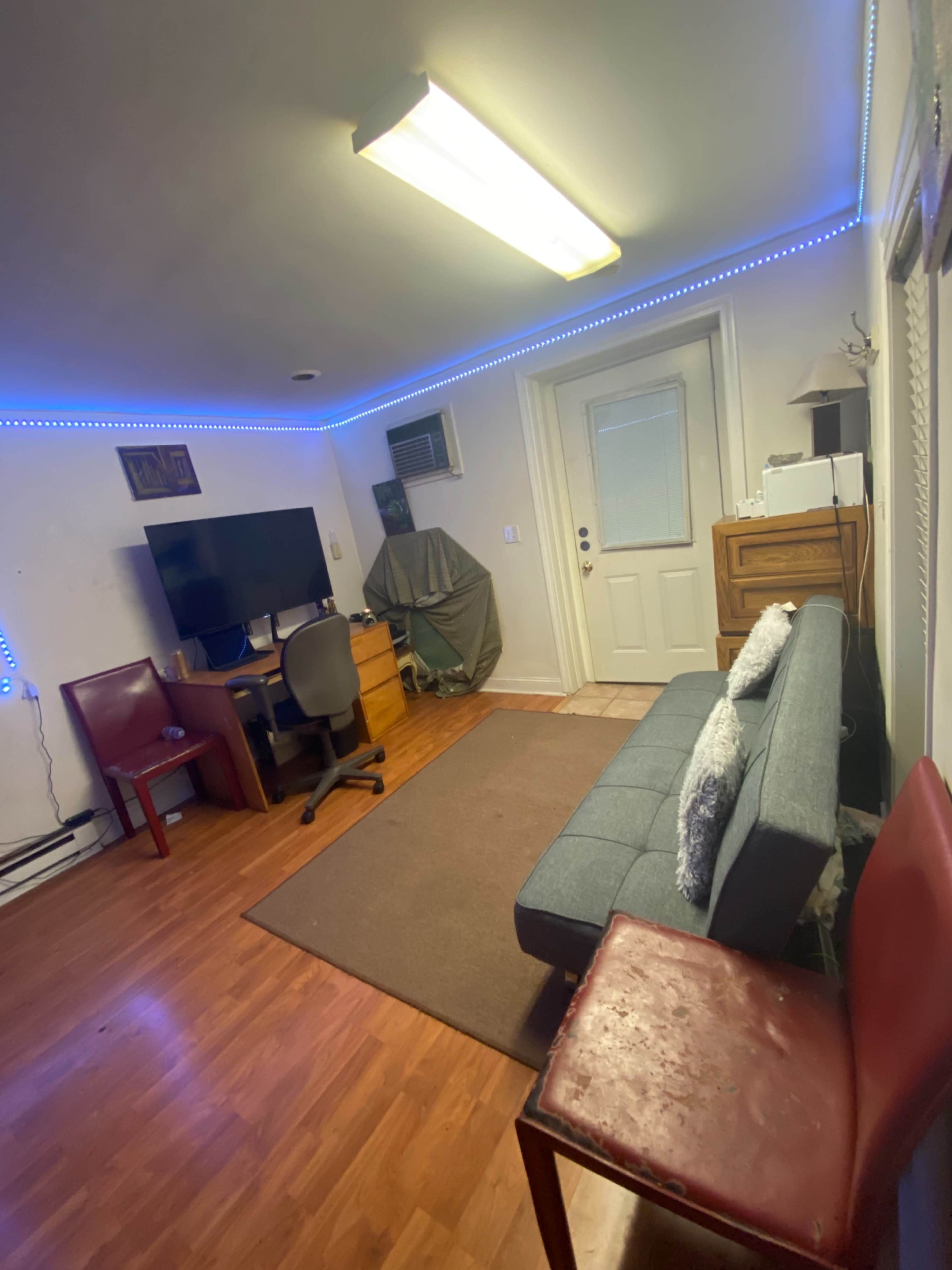 The image shows a small room with a gray sofa, a desk with a computer monitor, and two chairs, illuminated by blue LED light strips along the ceiling.