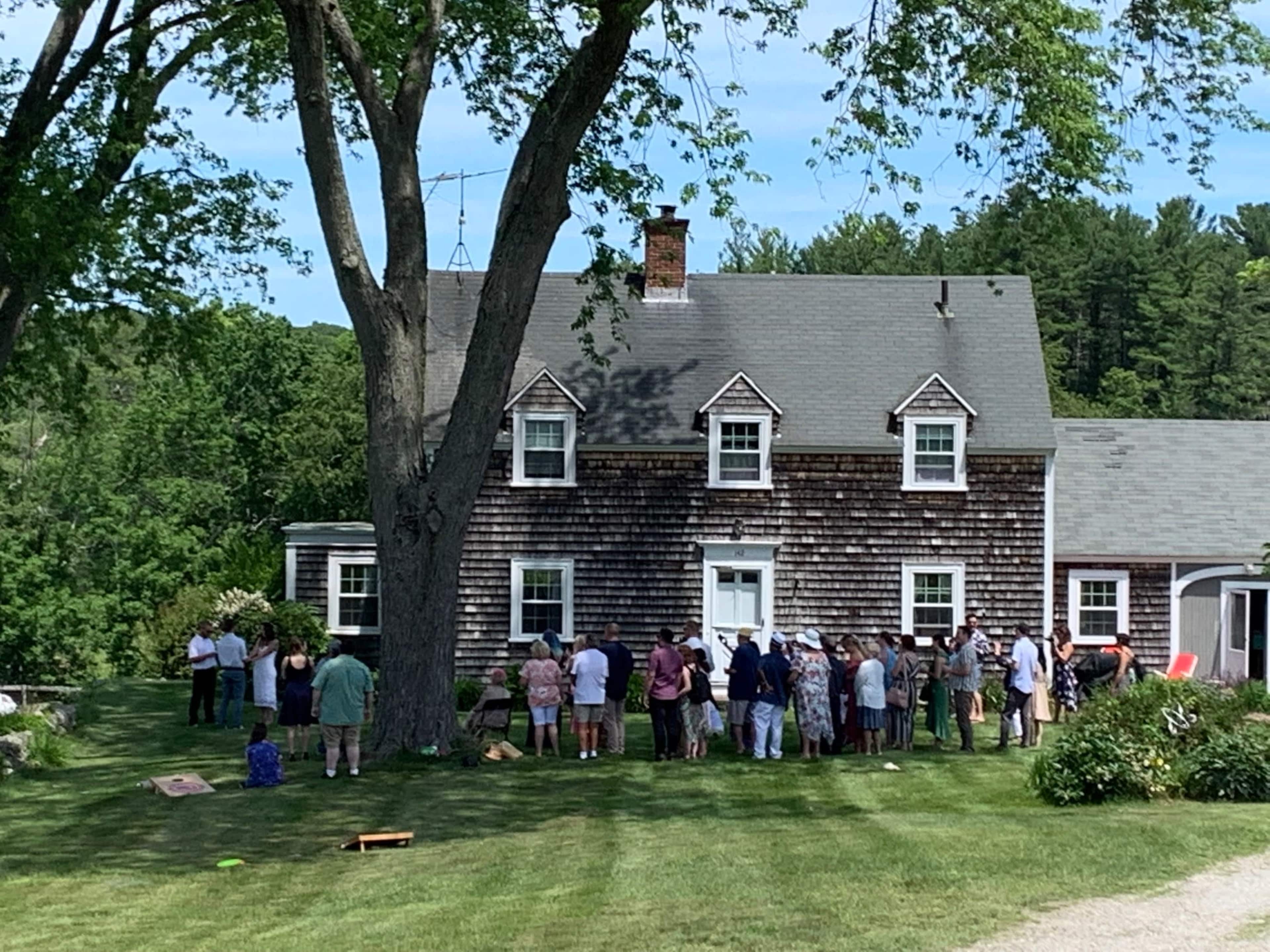 A large group of people gathers on the lawn near a two-story wooden house surrounded by trees.