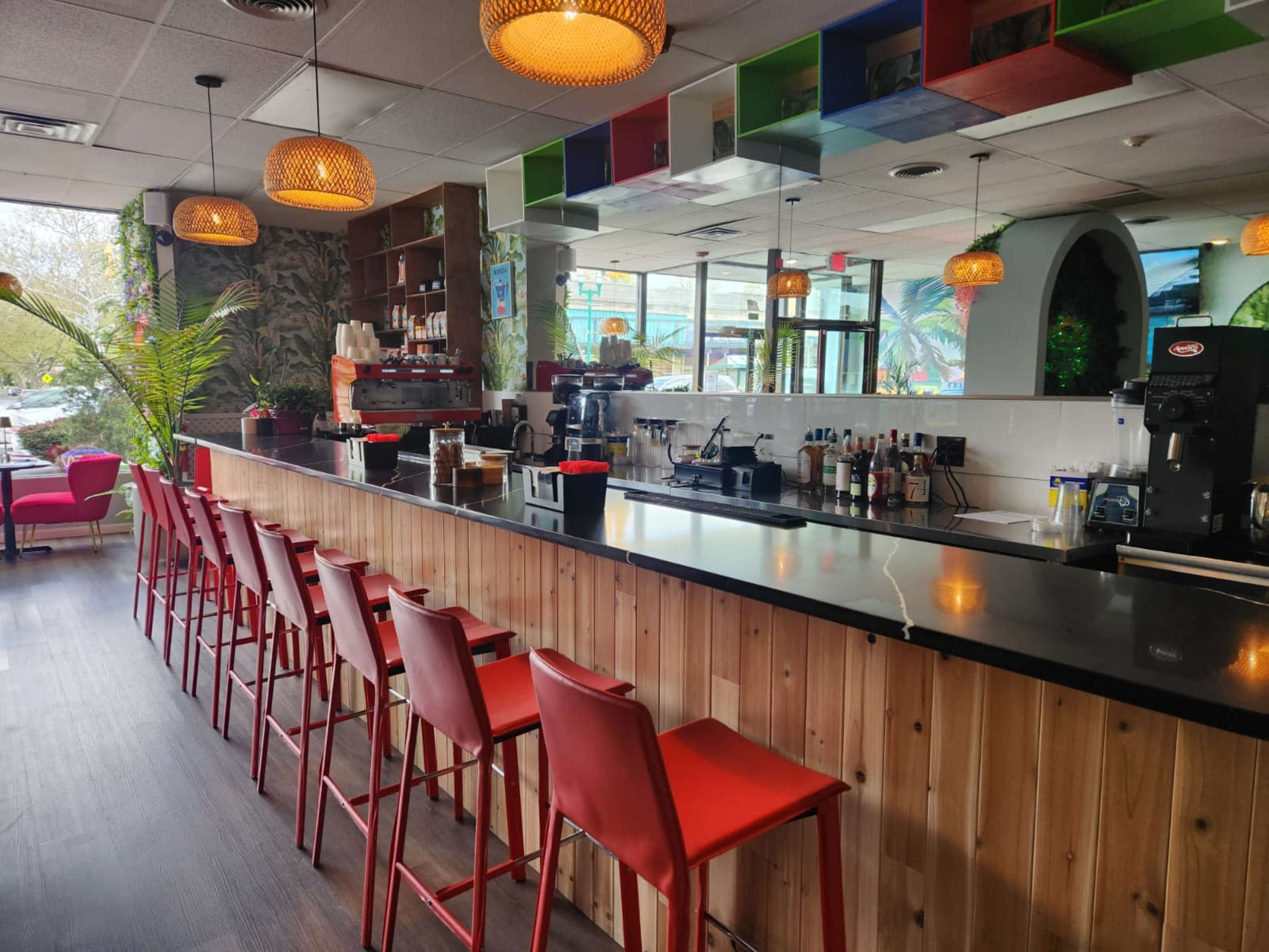 The image shows a bar area with red stools lined up against a wooden counter, colorful pendant lights hanging from the ceiling, and a variety of beverages on display in a bright, modern café setting.
