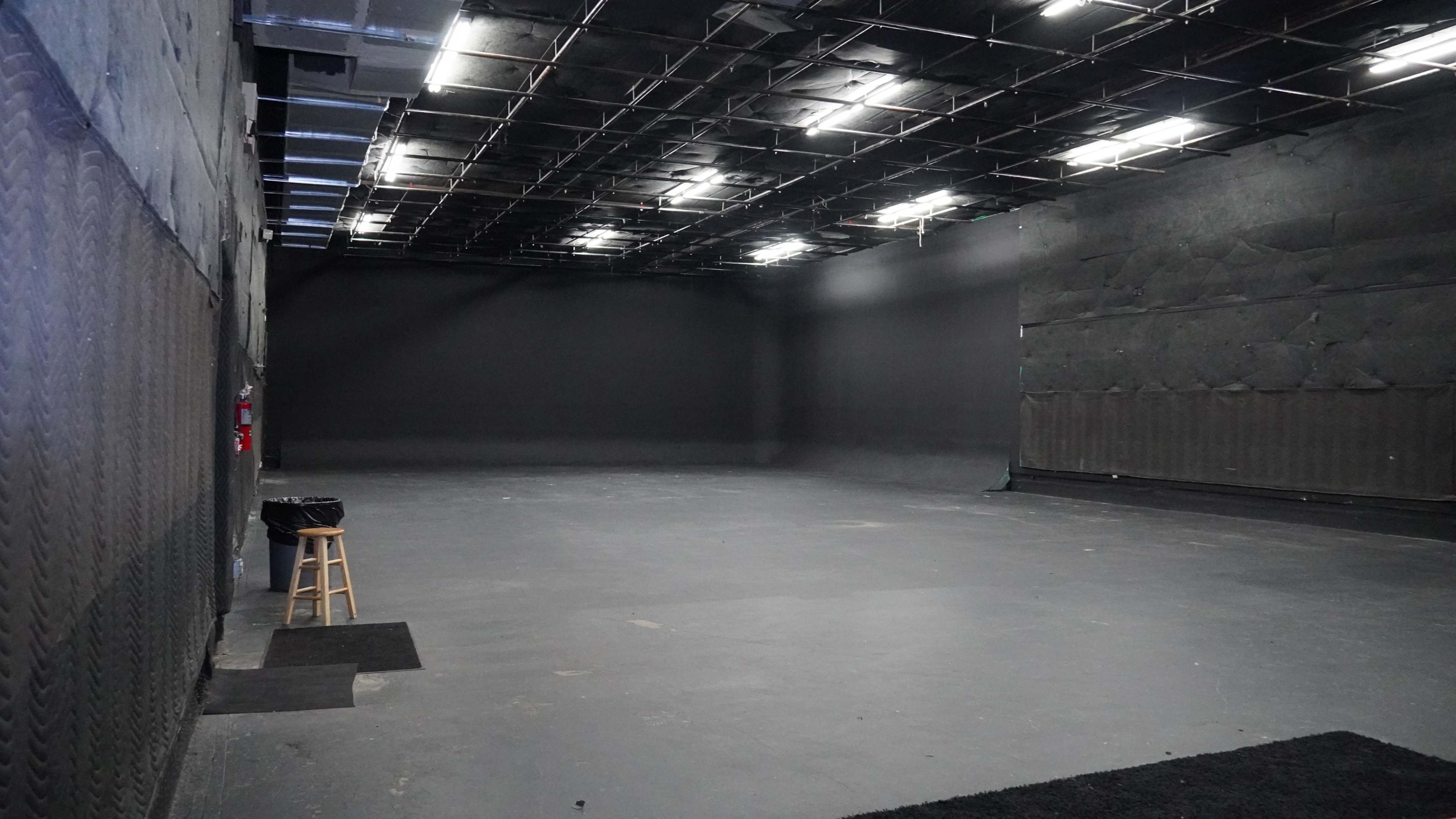The image shows a large, empty studio space with black walls and a concrete floor, featuring overhead lighting and minimal furnishings.