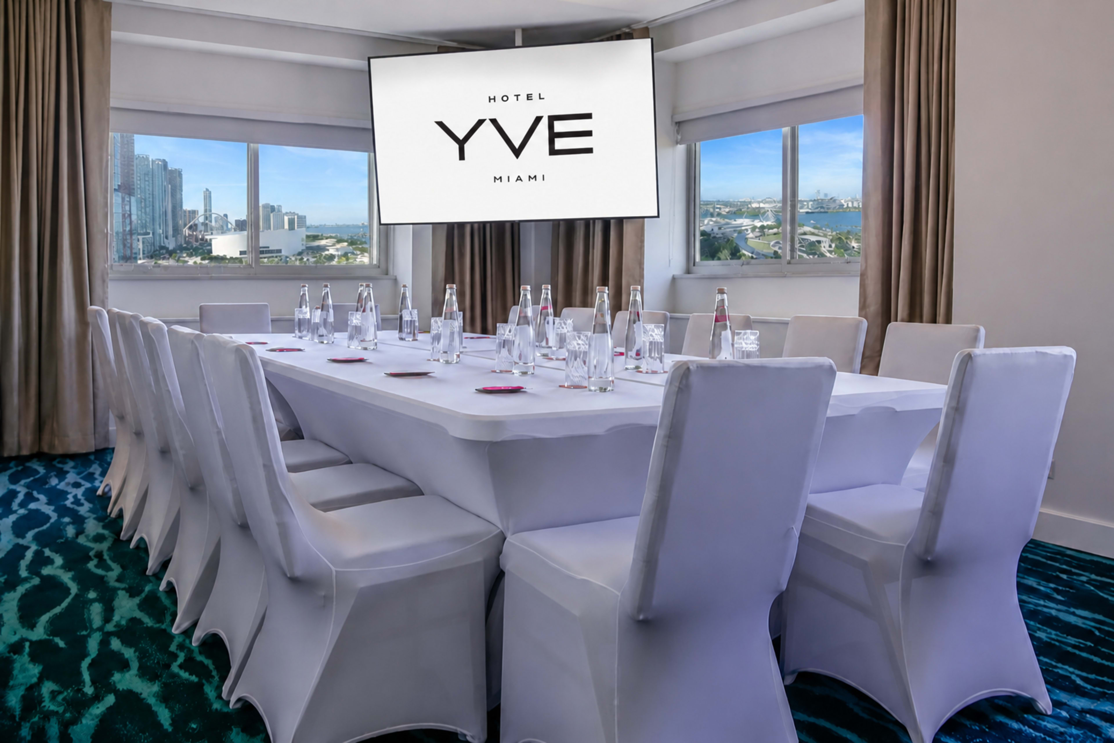 A conference room at Hotel YVE Miami features a long table set for a meeting, with a large screen displaying the hotel's logo and a view of the city outside.