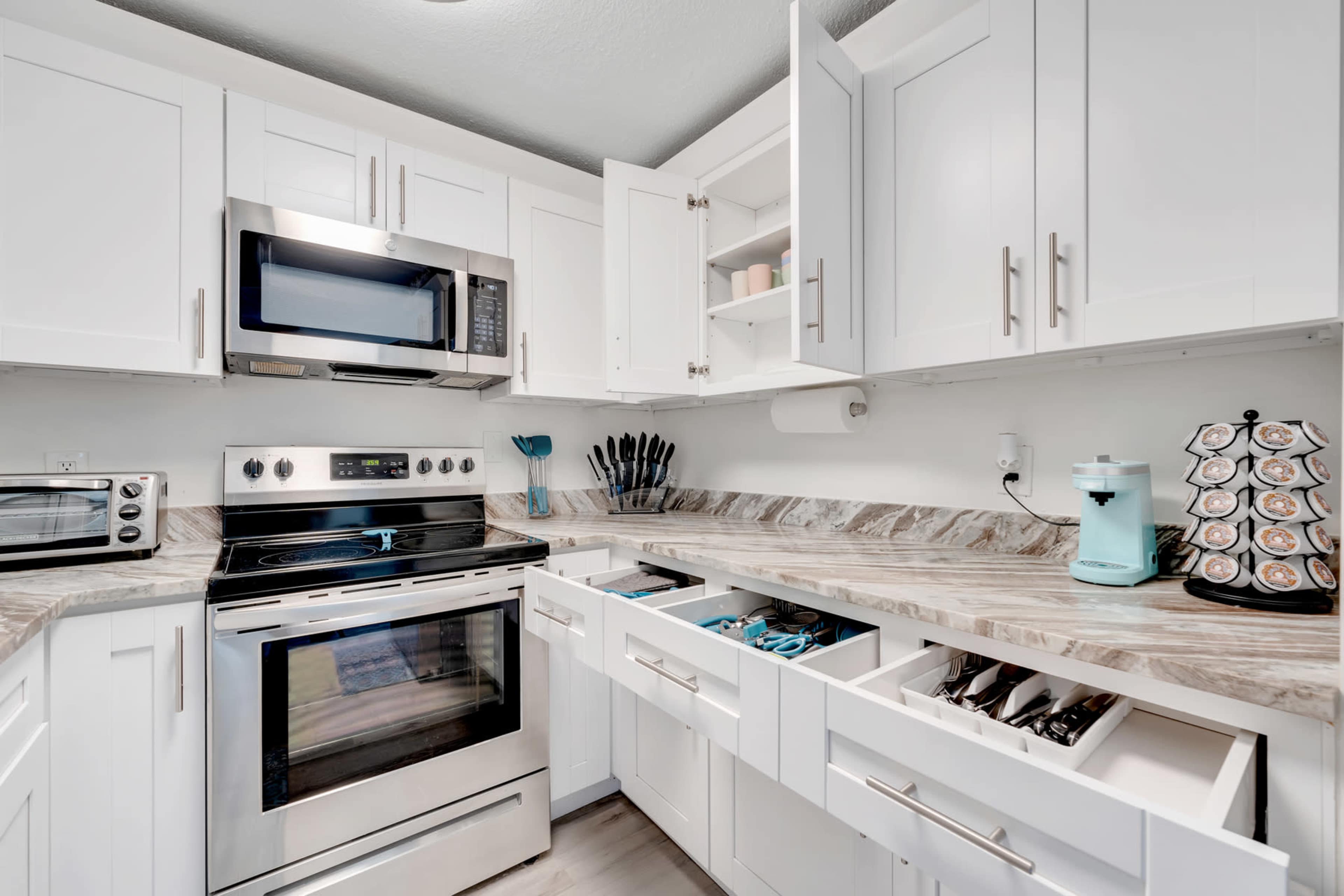 A modern kitchen features white cabinets, stainless steel appliances, and a marble countertop with organized drawers.