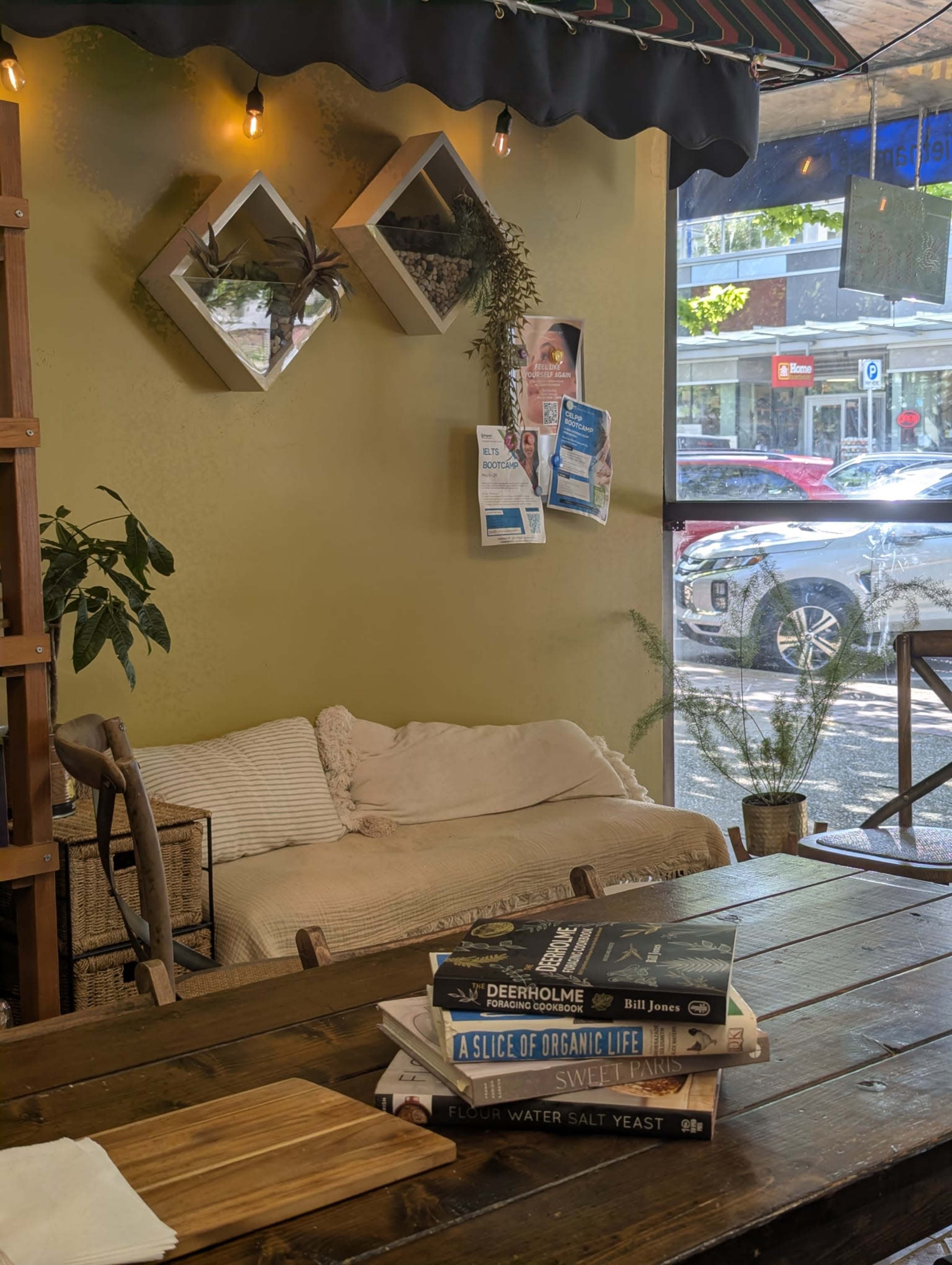 The image shows a cozy café interior featuring a wooden table, a couch against the wall, and books stacked on the table, with a window displaying a street view outside.