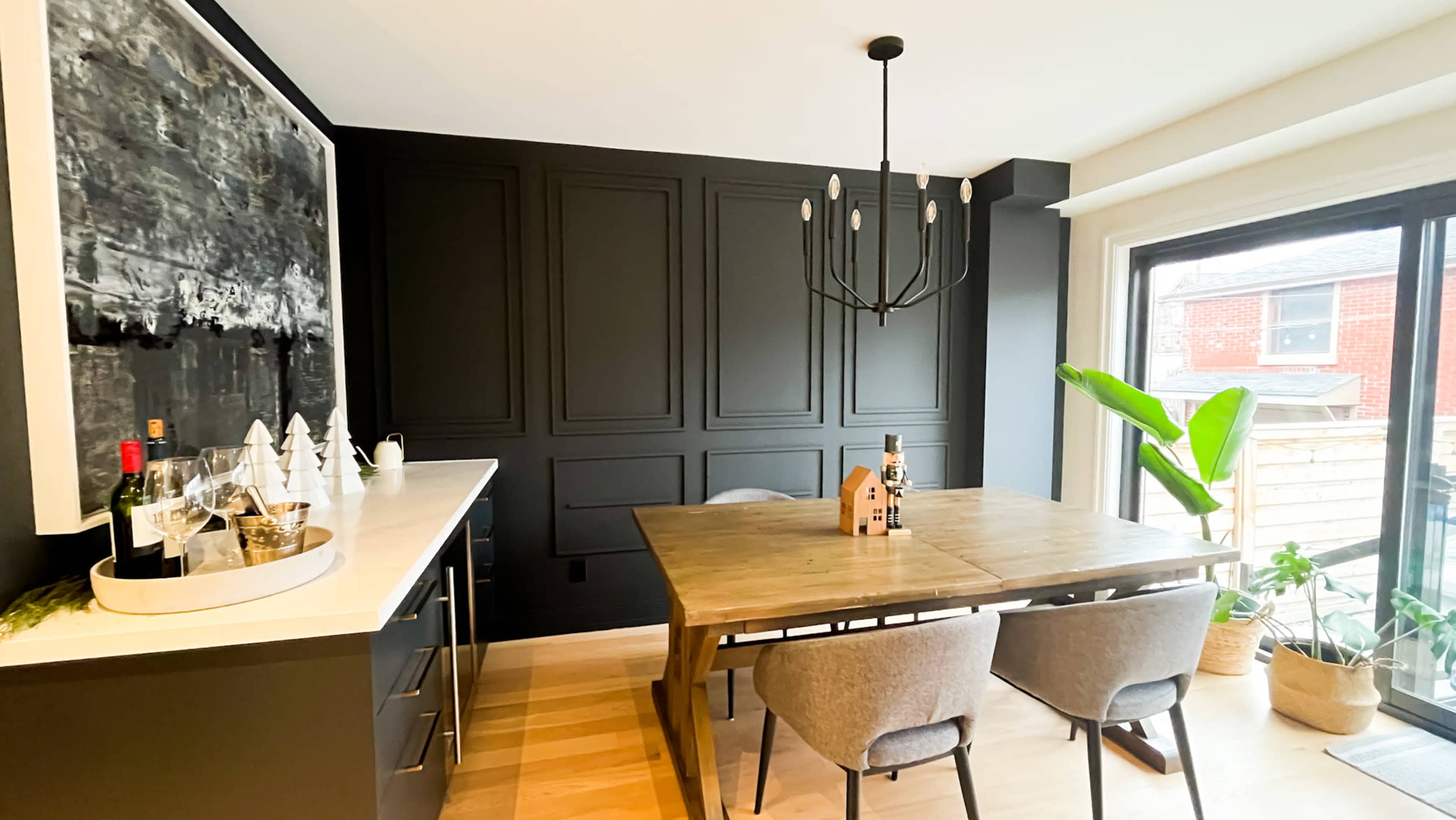 The dining area features a wooden table surrounded by gray chairs, set against dark paneled walls and a large artwork, with a bar cart to the side and a window offering natural light.
