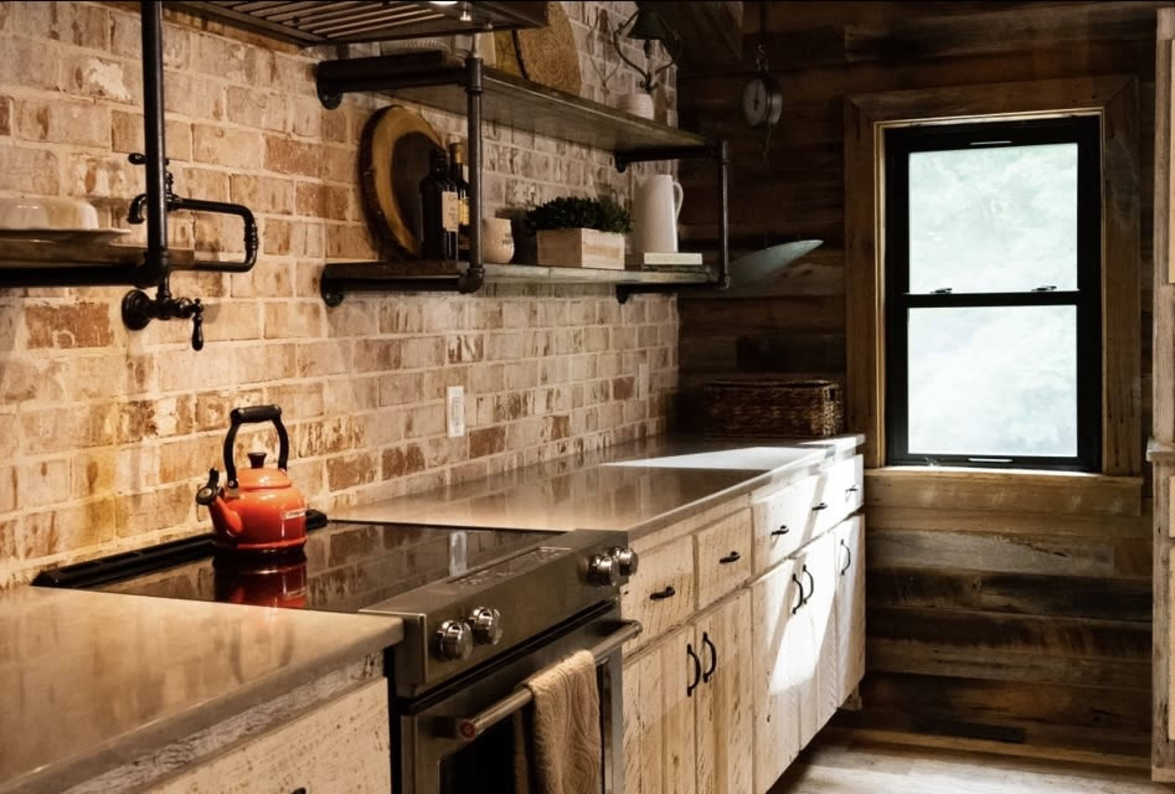 A kitchen with a brick wall, metal shelves, a stainless steel countertop, and a red kettle on the stove.
