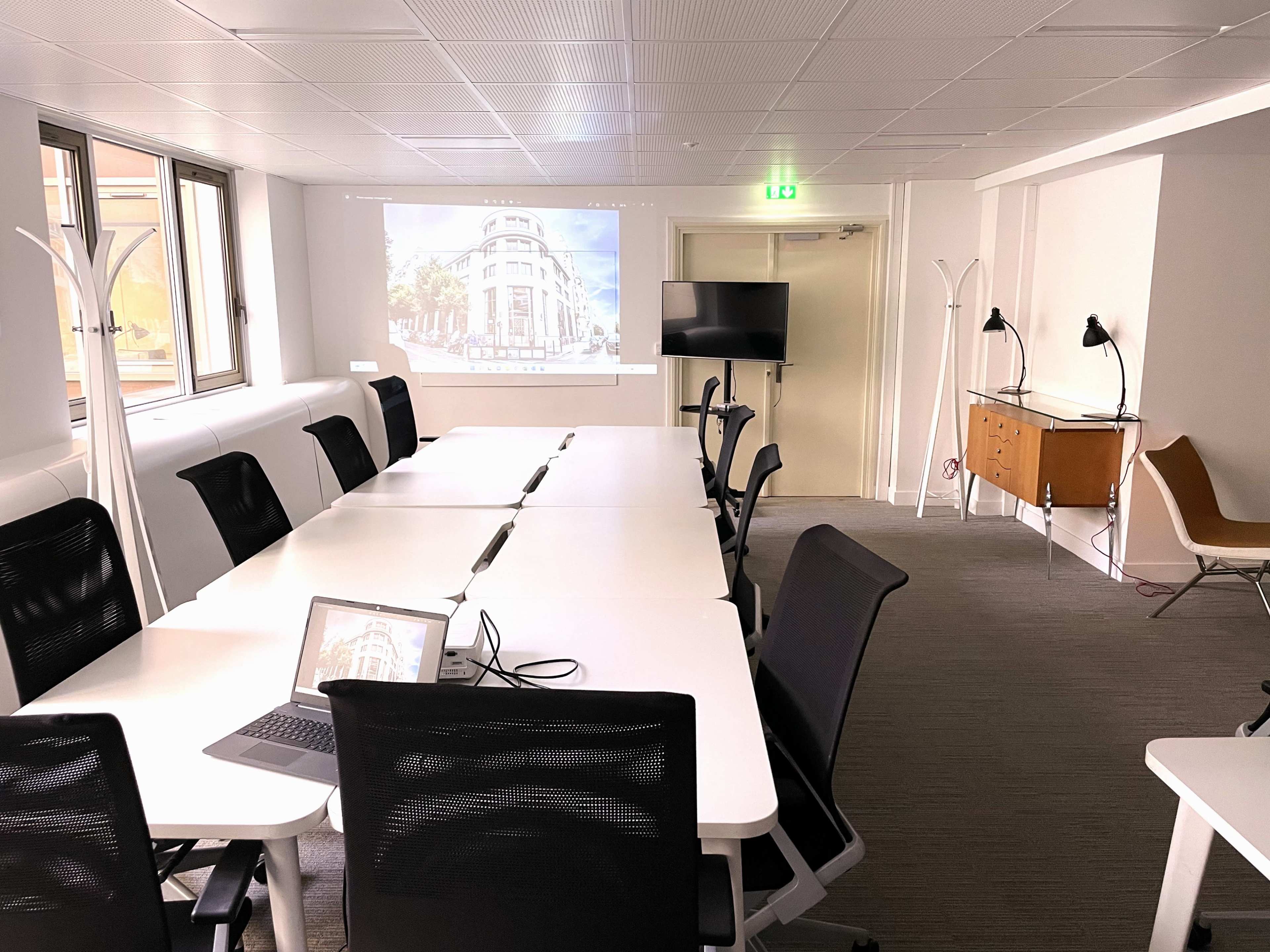 A modern conference room features a long table with chairs, a large screen displaying an image, a laptop, and various lighting fixtures.