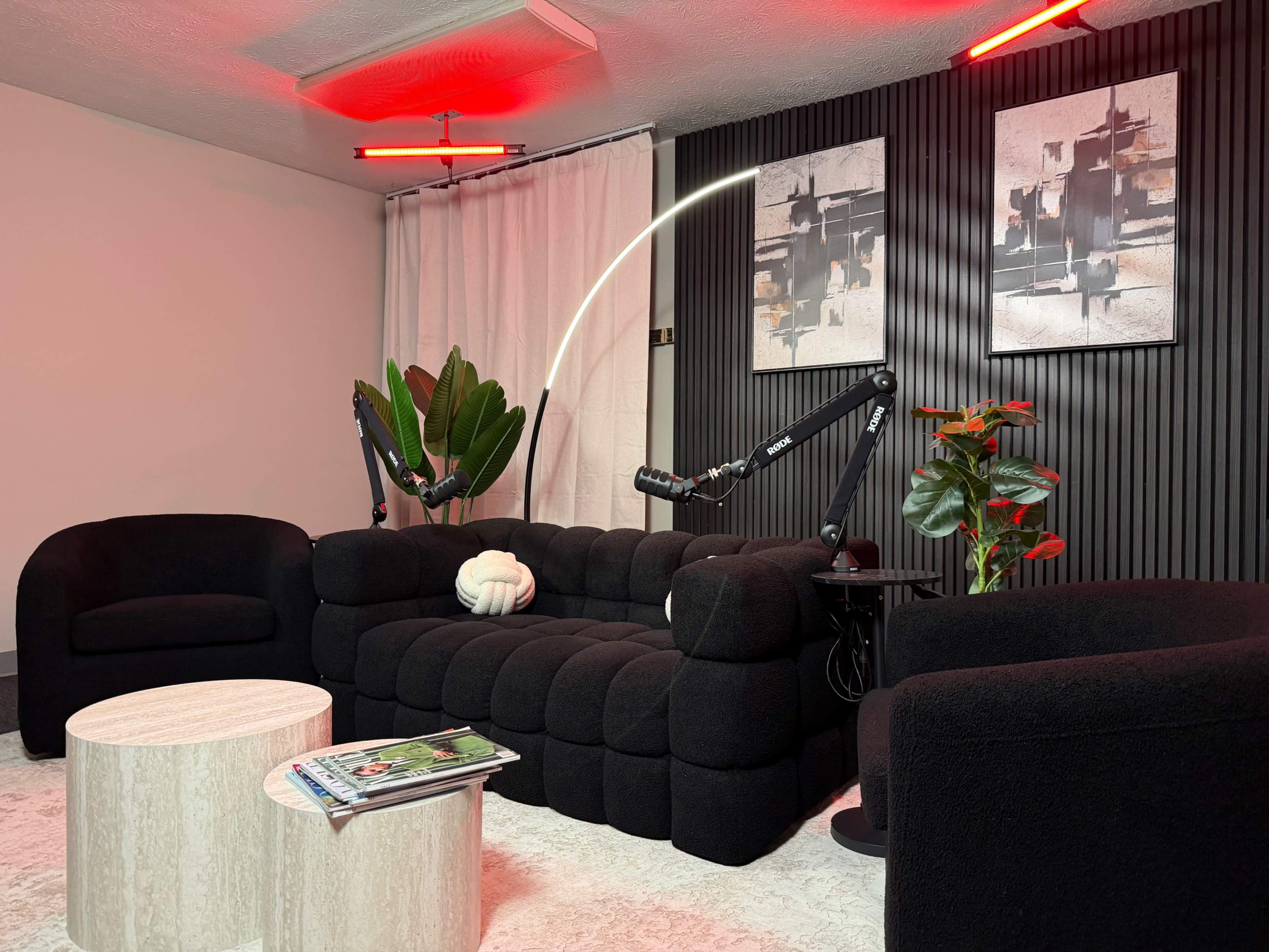 The image features a modern living room with a black sofa, two armchairs, a round coffee table, and decorative plants, alongside abstract wall art and red light fixtures.