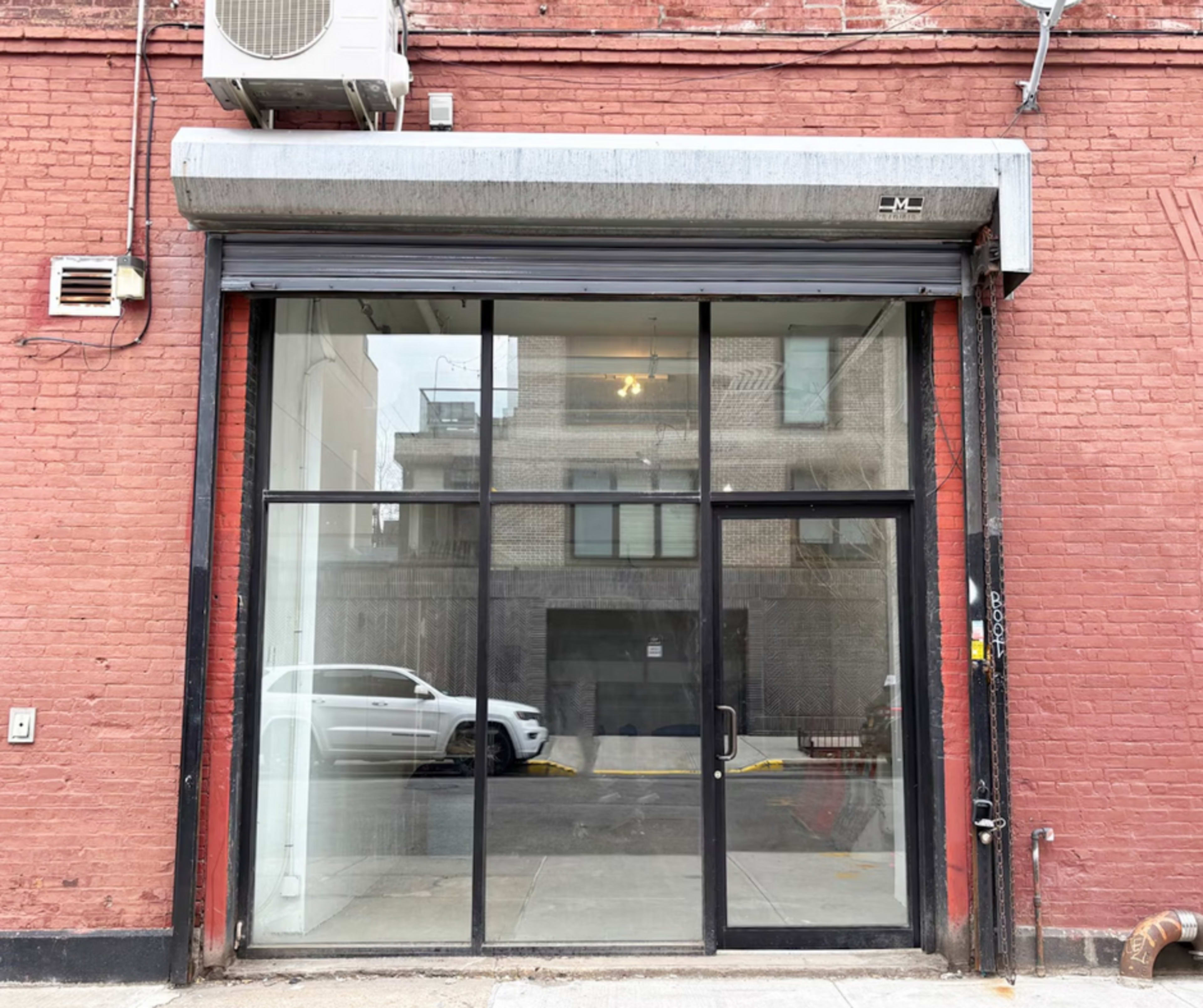 The image shows a vacant storefront with large glass panels and a metal awning, located on a brick wall.