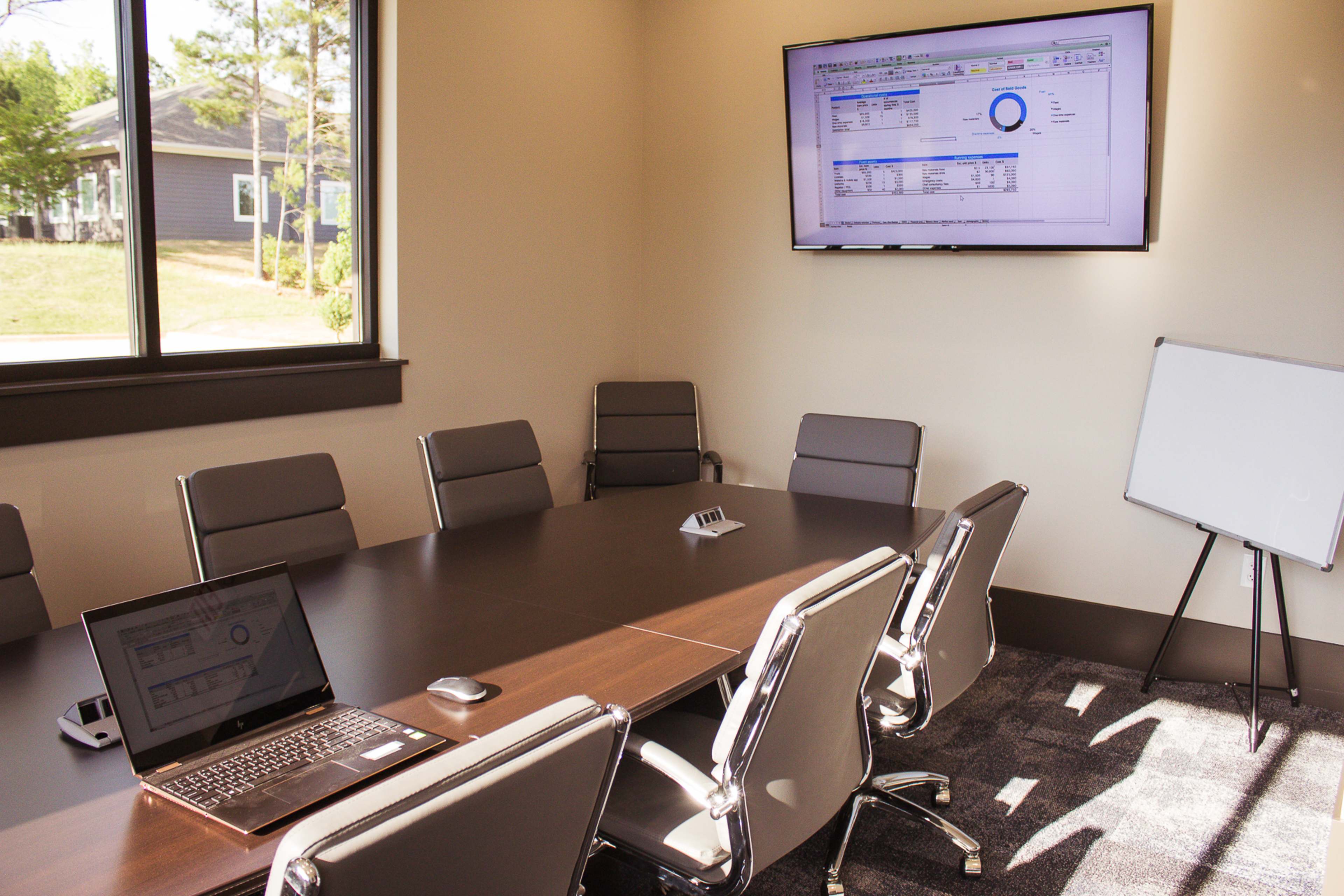 A modern conference room features a large table, eight chairs, a laptop on the table, a presentation screen on the wall, and a whiteboard.