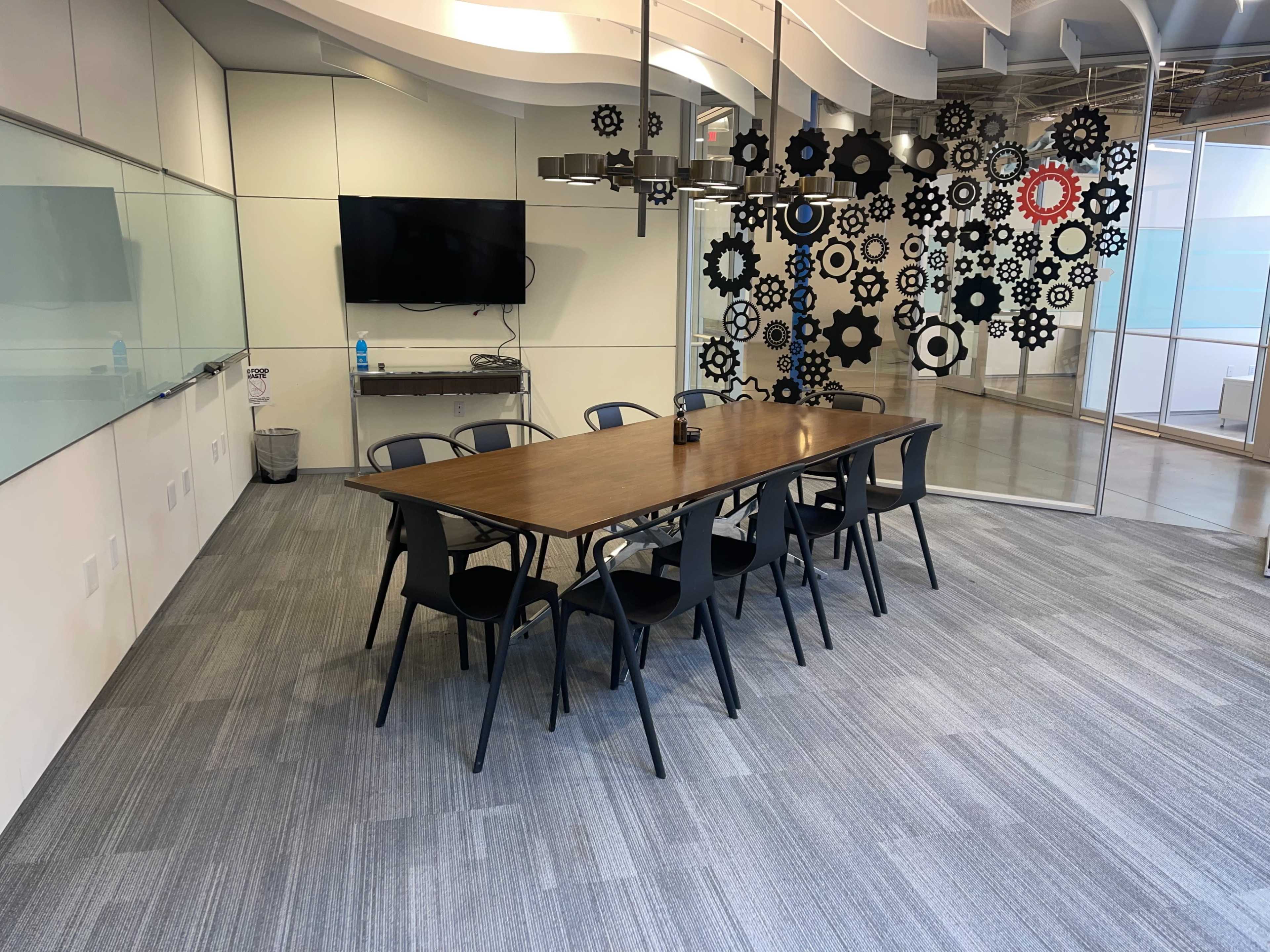 A meeting room features a long wooden table surrounded by black chairs, with a television on one wall and decorative gear shapes on another.