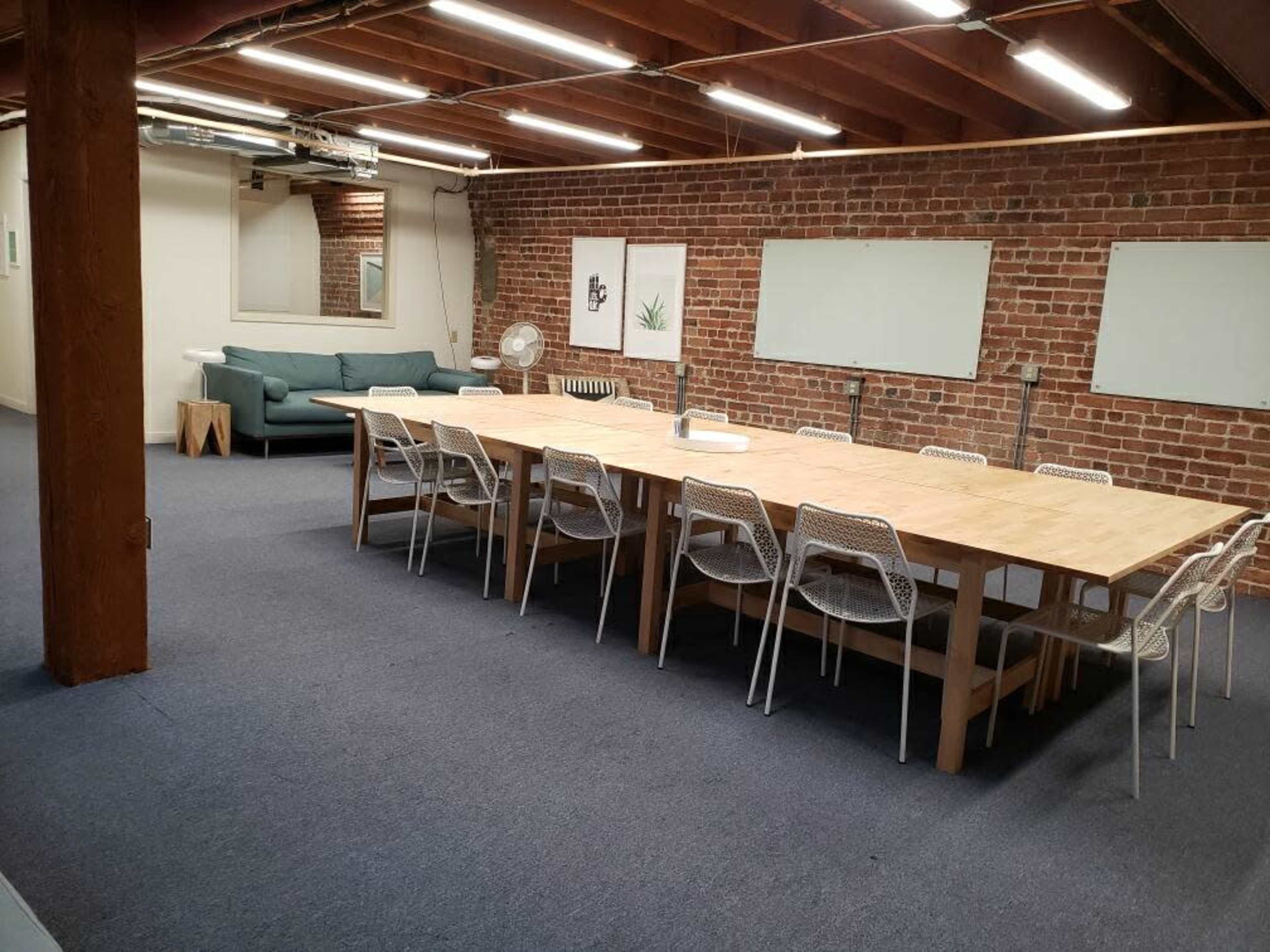 A spacious meeting room with a long wooden table surrounded by white chairs, a green couch in the background, and exposed brick walls.
