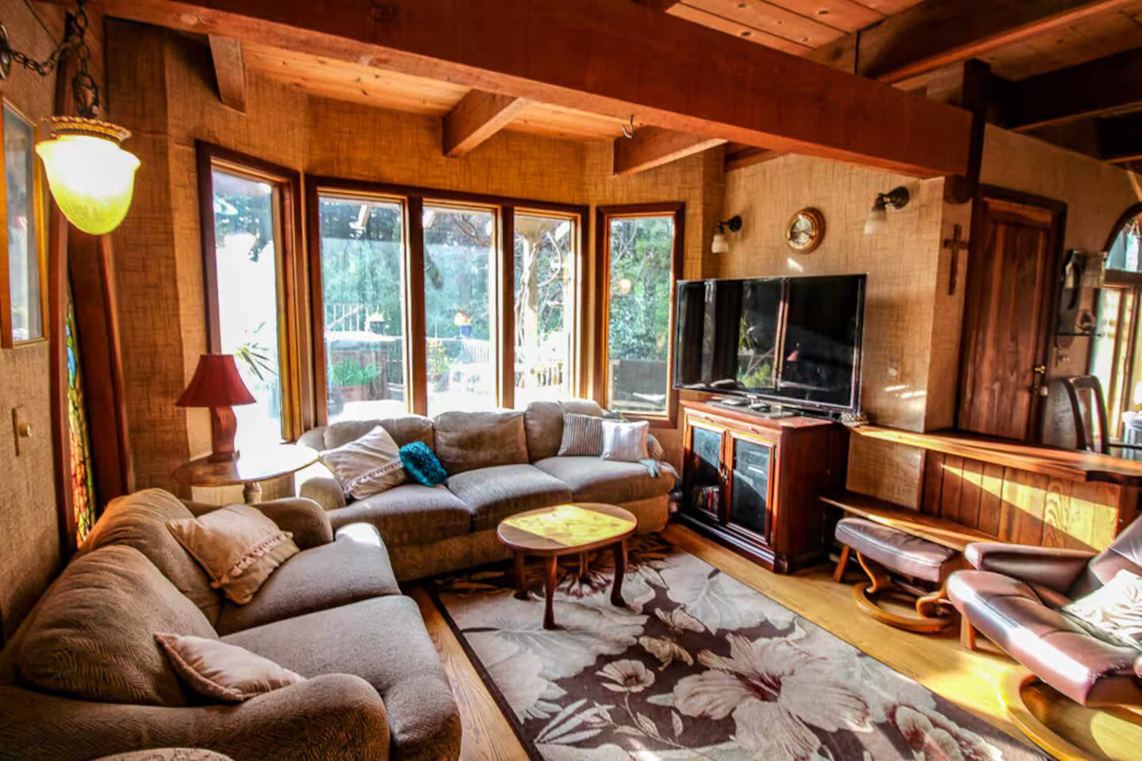 The image shows a cozy living room with a large sectional sofa, a round coffee table, and a flat-screen TV, surrounded by multiple windows that let in natural light.