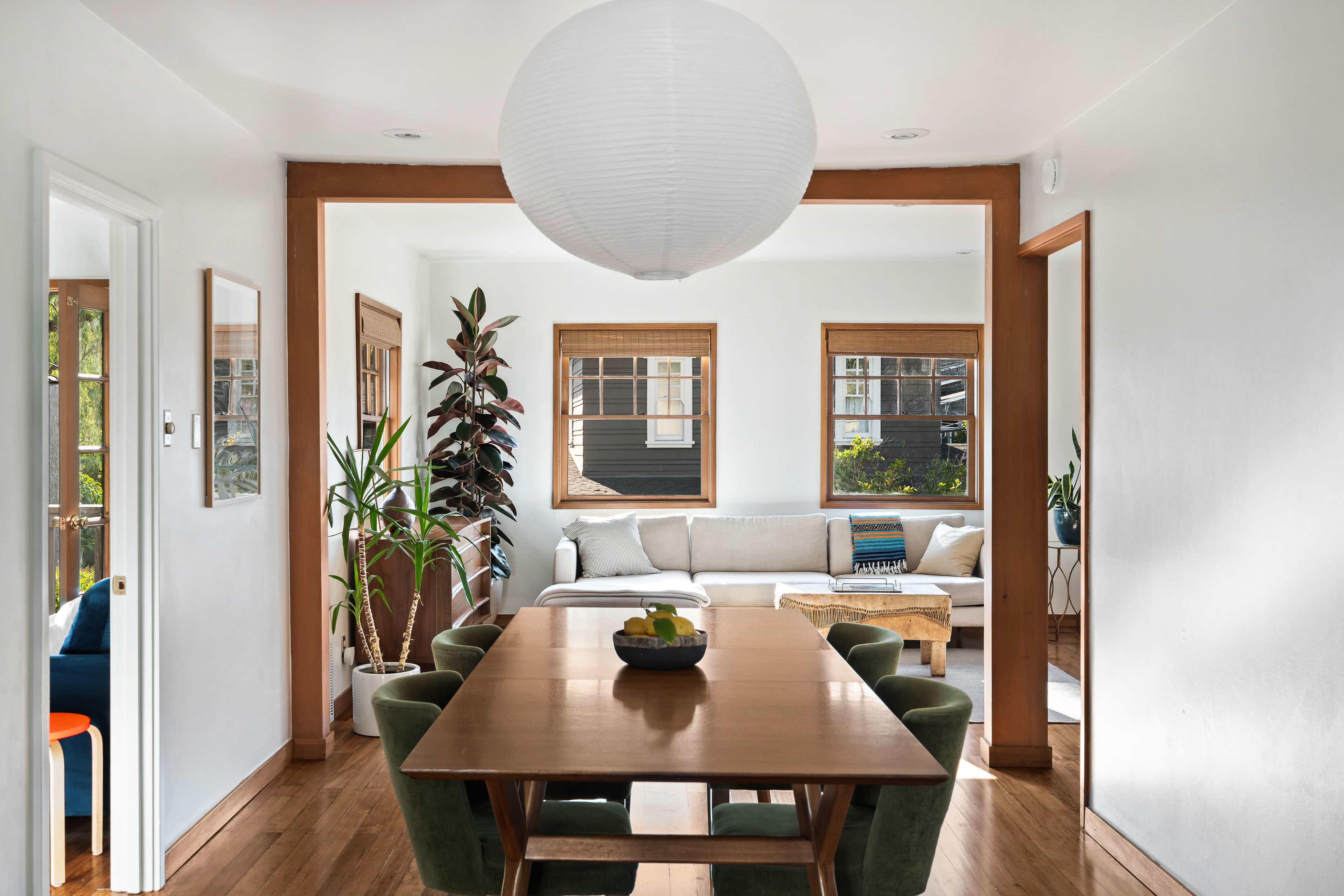 A dining area featuring a wooden table with green chairs, surrounded by plants and natural light from large windows.