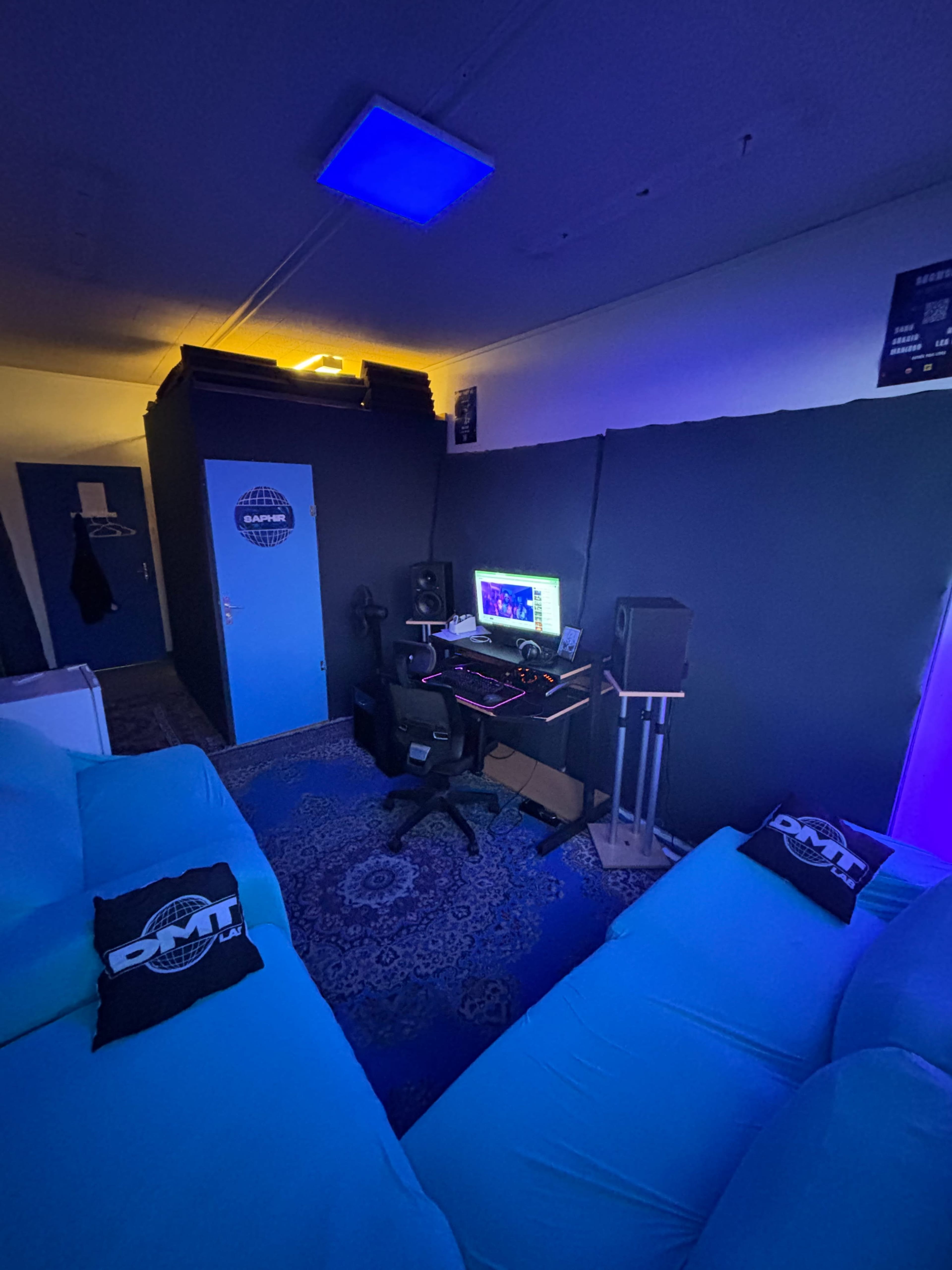 A cozy music studio featuring a computer setup with monitors, mixing equipment, and blue lighting, surrounded by dark walls and a comfortable seating area.