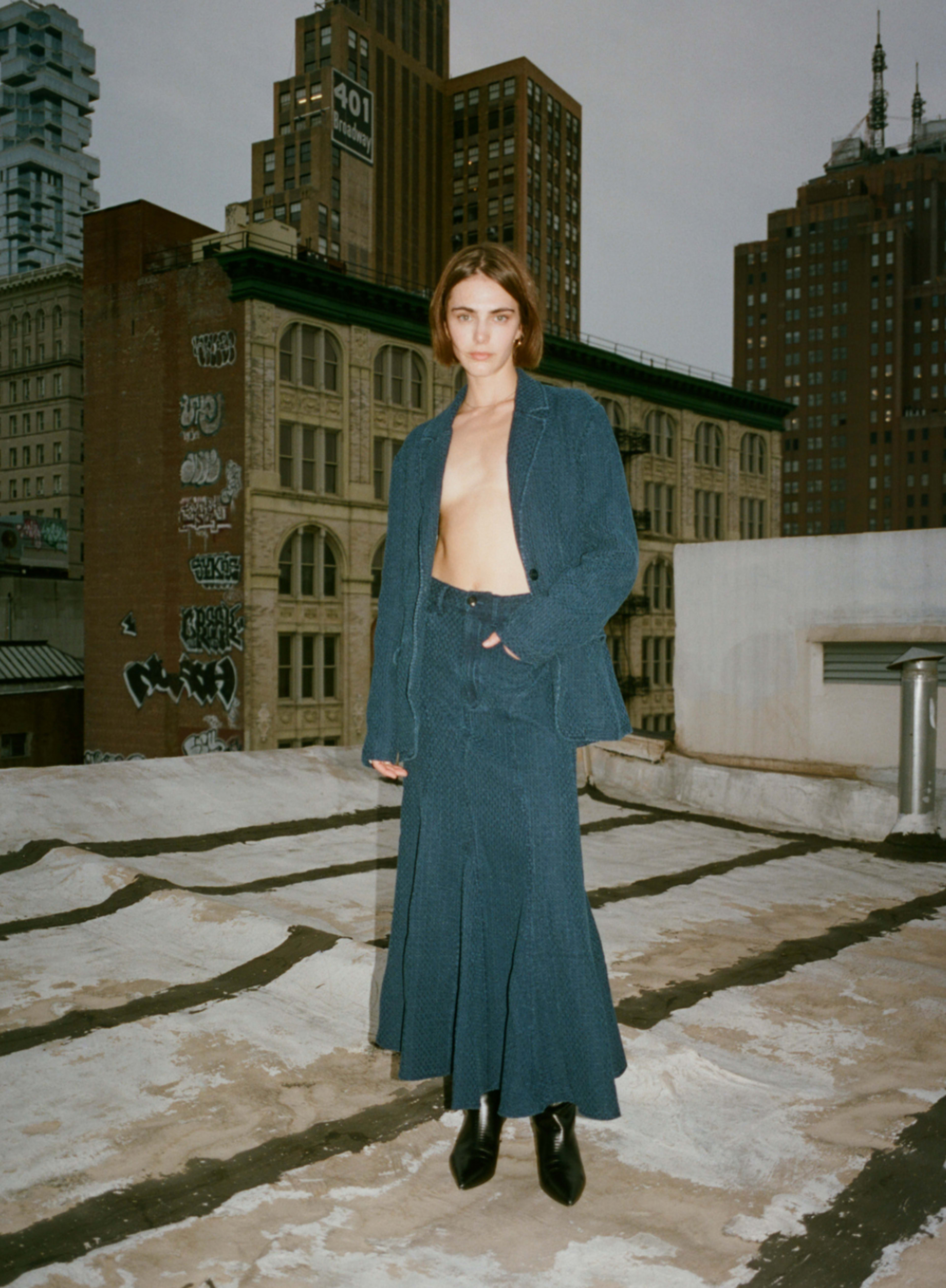A model stands on a rooftop wearing a blue two-piece outfit, with tall buildings and graffiti in the background.