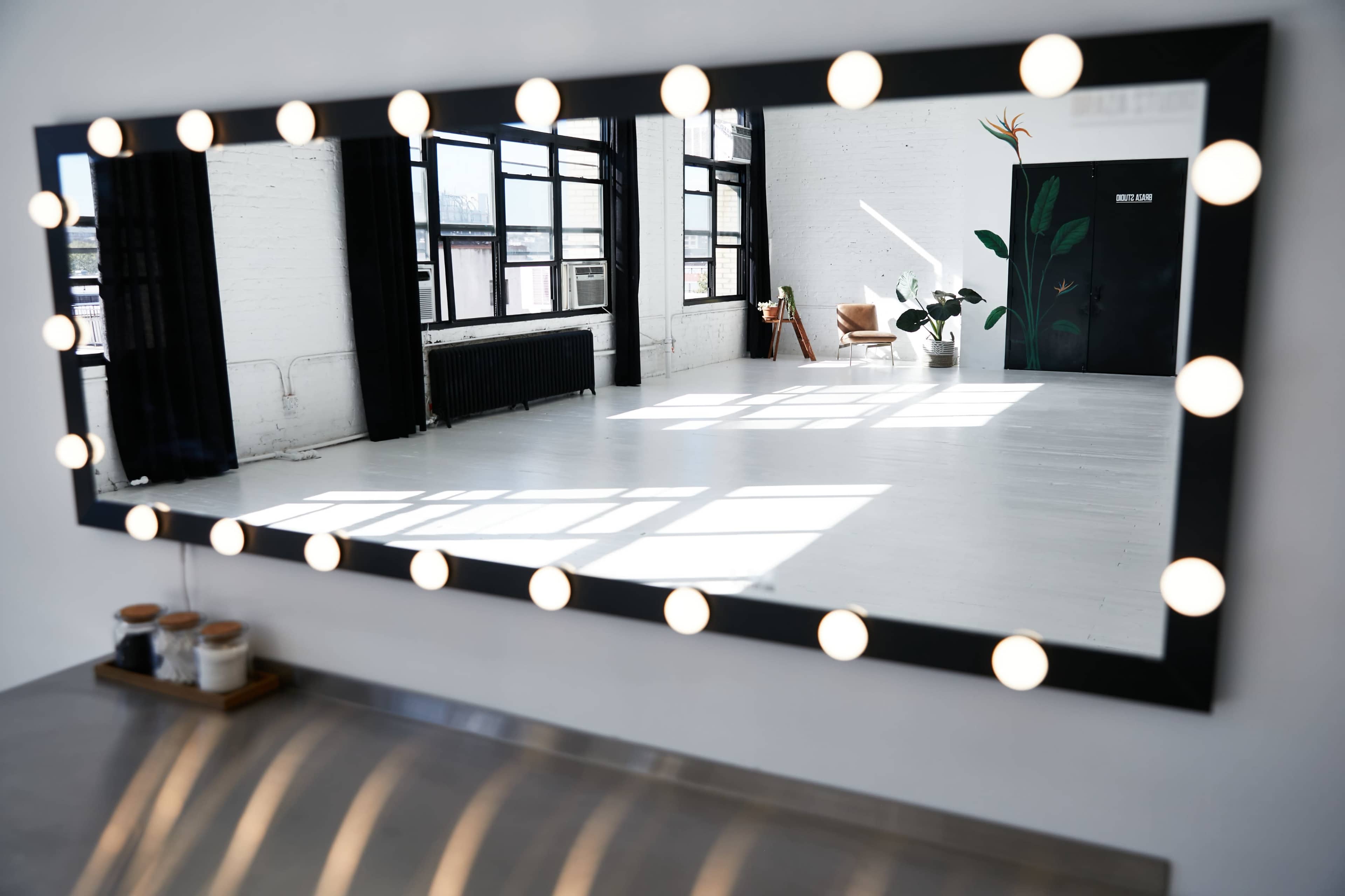 A large mirror framed with lights reflects a bright room featuring windows, a potted plant, and minimalist furniture.