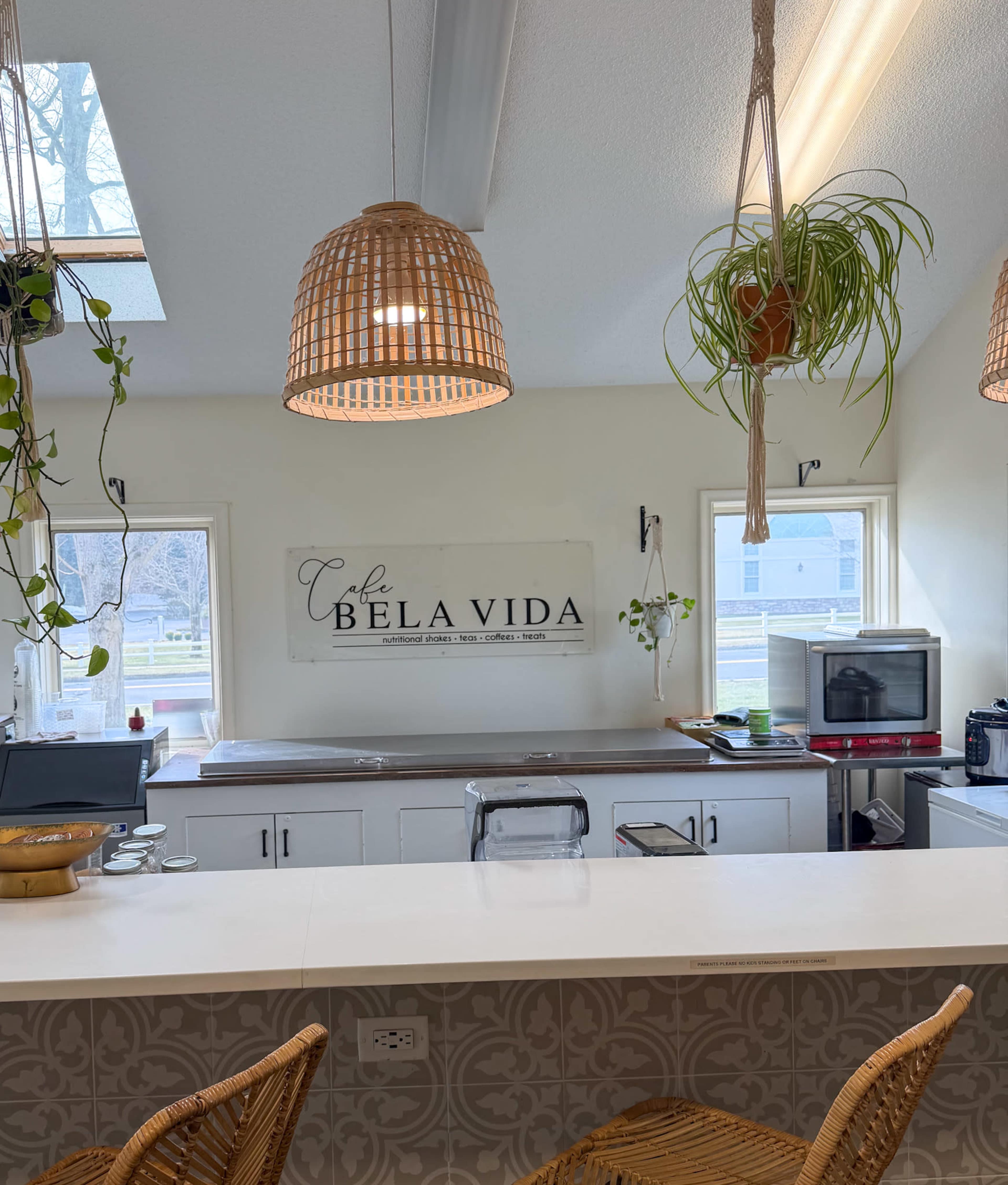 The image shows a cozy café interior with a serving counter, hanging rattan light fixtures, and indoor plants.