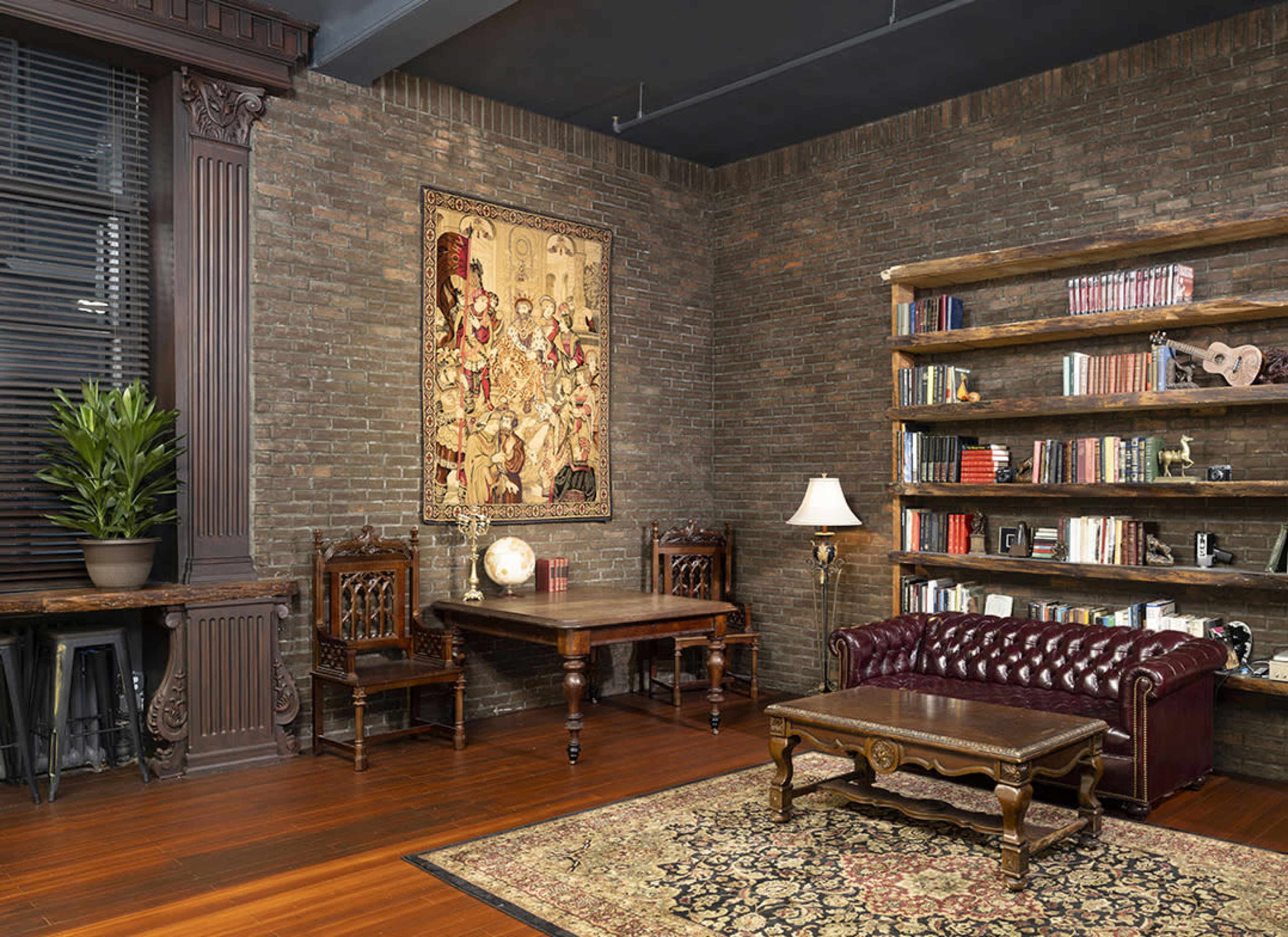 A cozy room with a brick wall, a dark wooden table, two chairs, a leather sofa, a bookshelf filled with books, and a decorative rug on the wooden floor.
