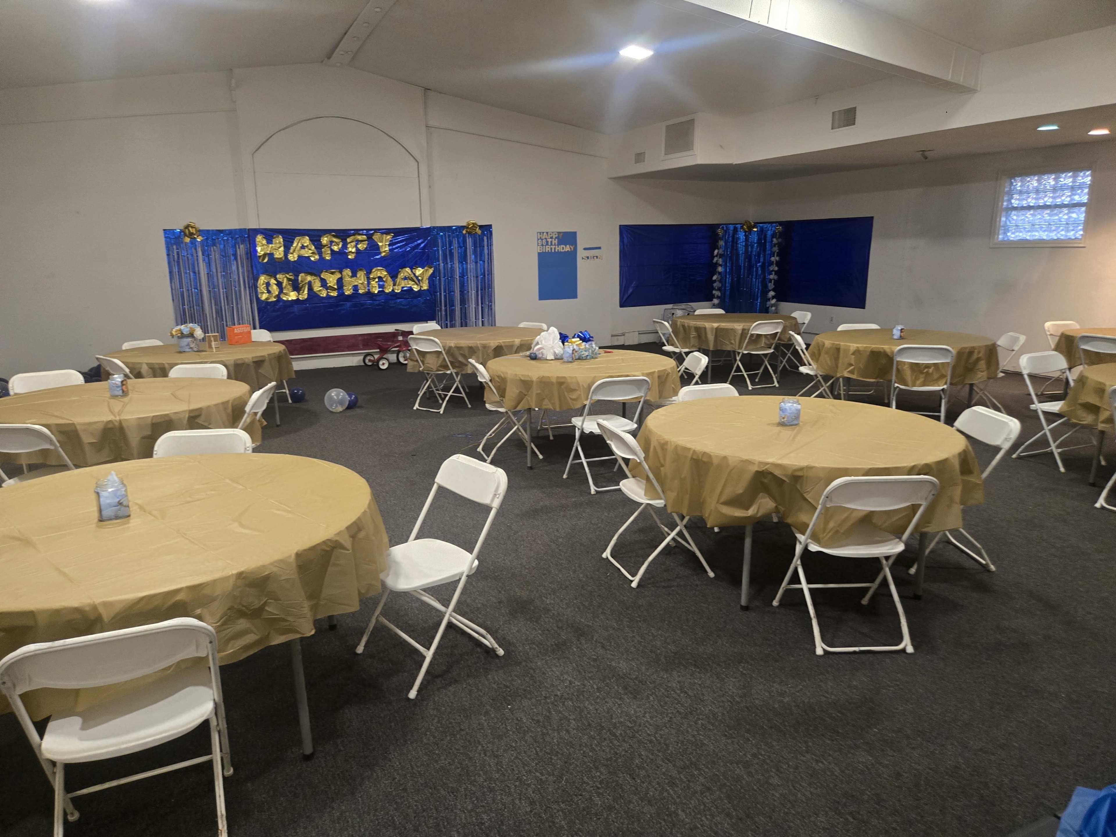 The image shows a decorated event space with circular tables covered in gold tablecloths, arranged around the room, and a festive backdrop that reads "HAPPY OLNYDAY."