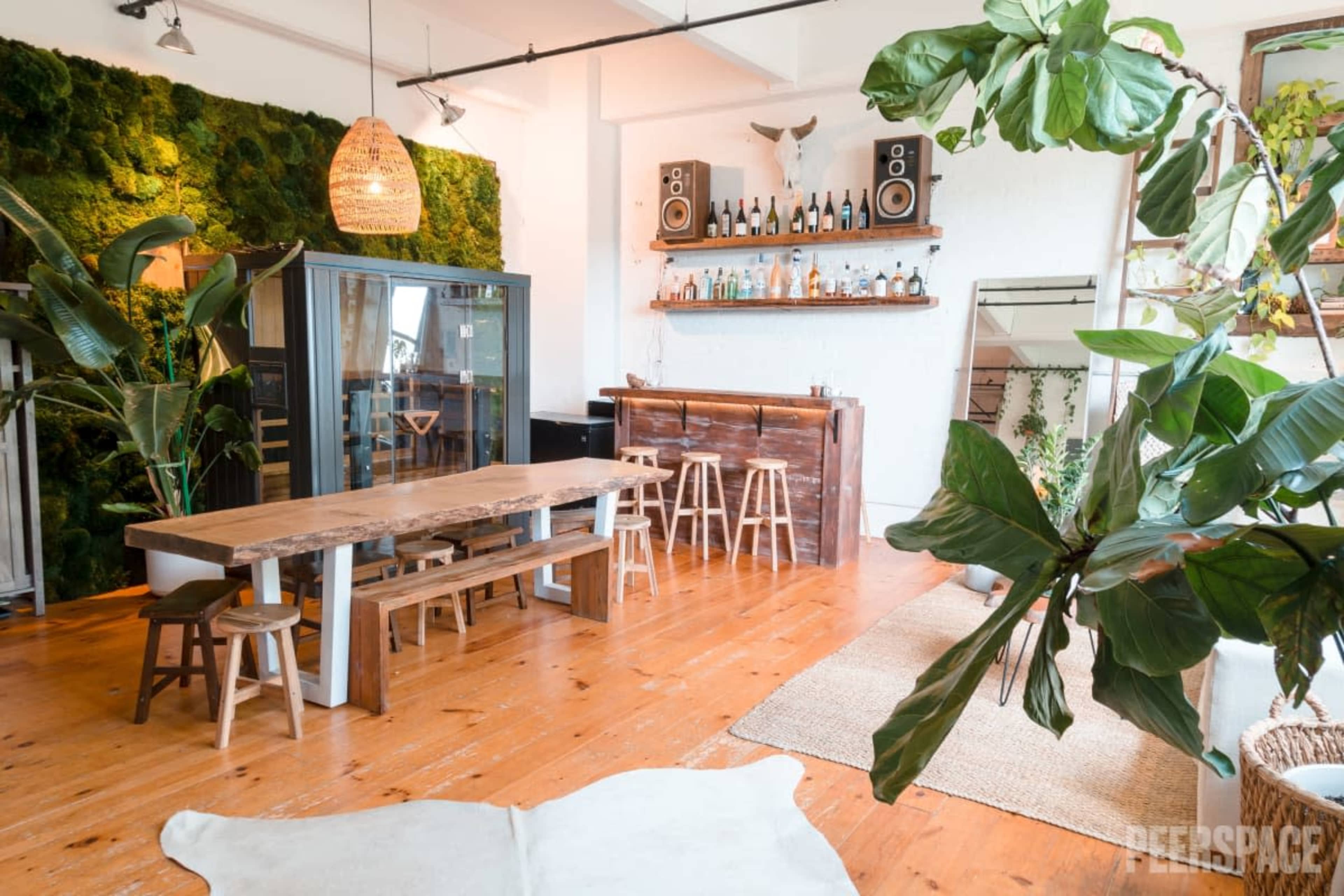 The image showcases a spacious interior featuring a large wooden dining table, bar area with drinks, and a green moss wall, surrounded by various plants.