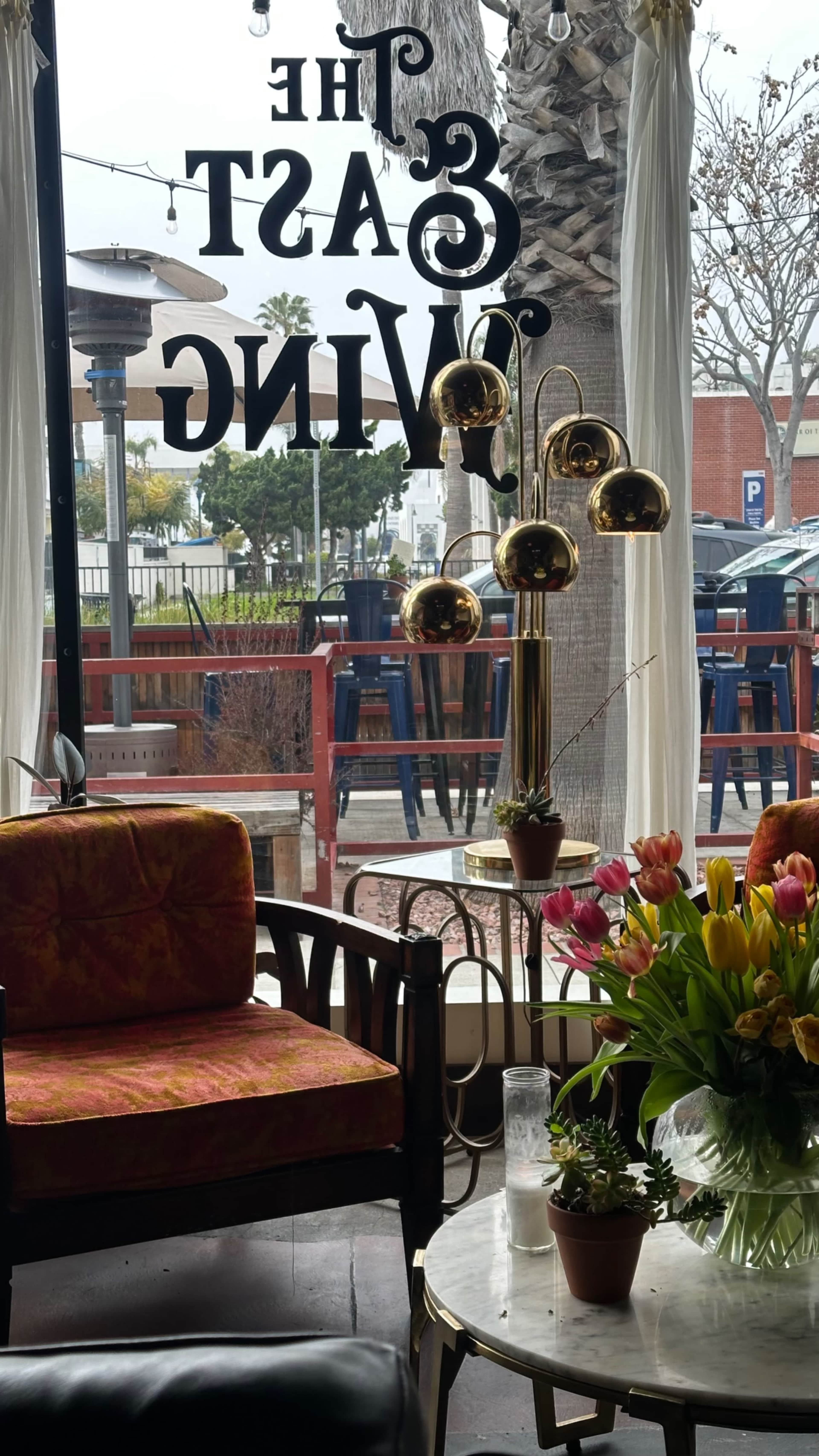 The image shows a cozy interior with a vibrant armchair, a gold floor lamp, and a bouquet of tulips on a coffee table, overlooking an outdoor seating area through a large window.