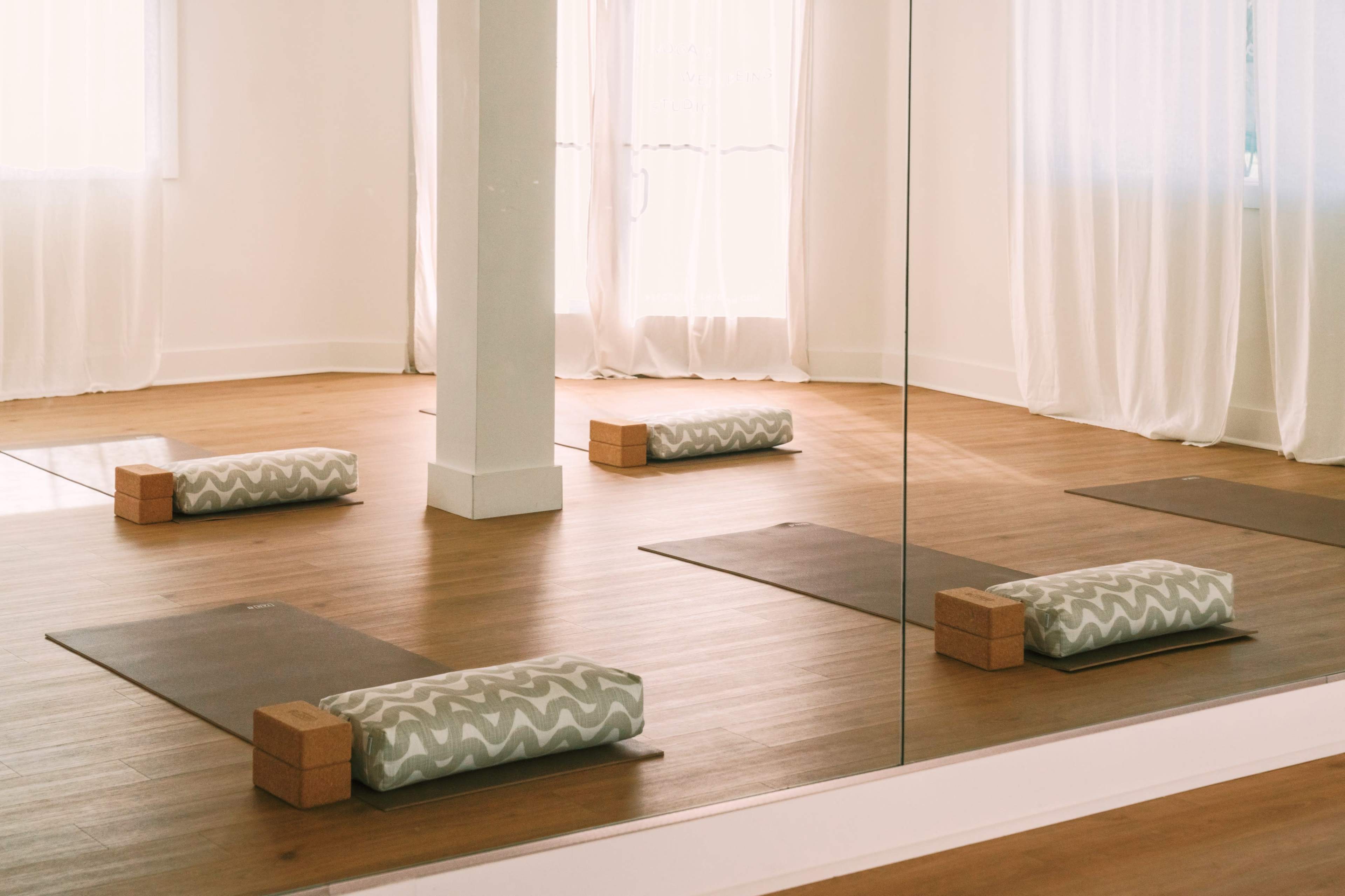 The image shows a serene yoga studio with wooden flooring, two yoga mats, yoga bolsters, and blocks arranged neatly in front of a mirror.