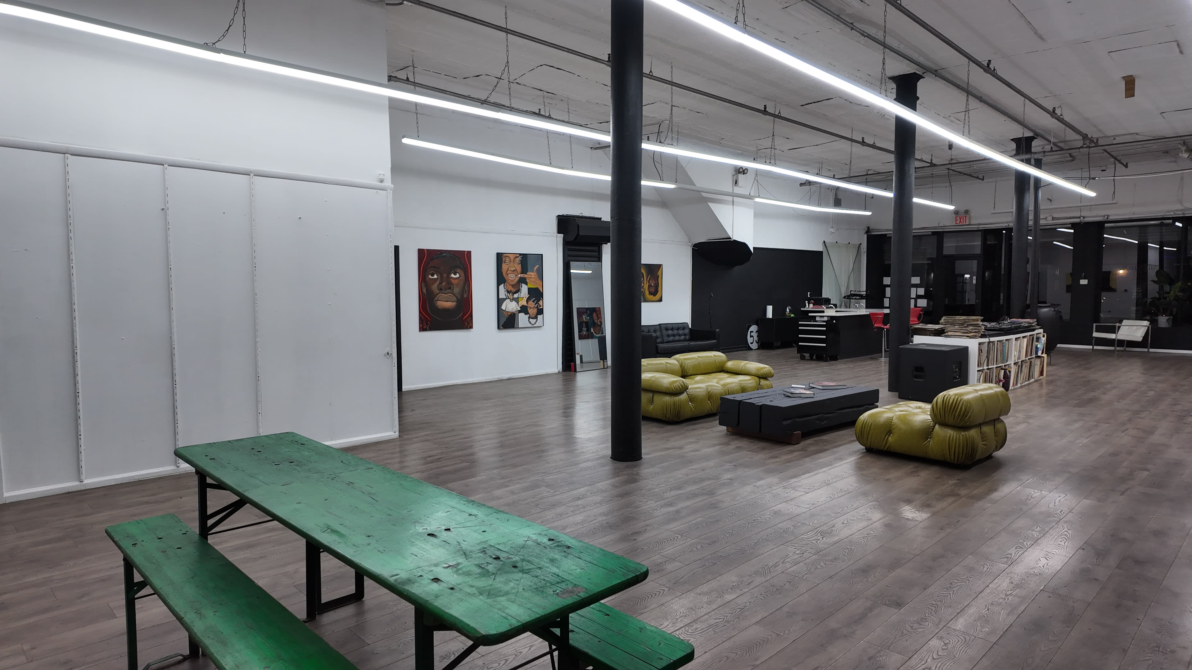 The image shows a spacious, modern interior with wooden flooring, featuring green picnic tables, yellow seating, and various artworks on the walls.