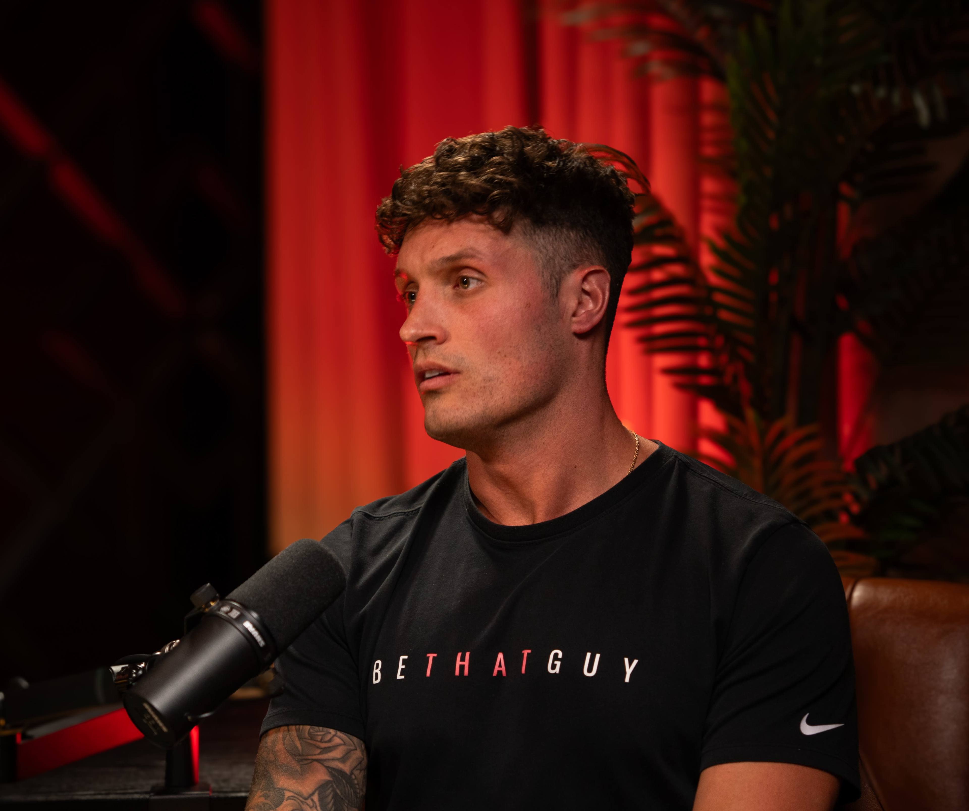 A man with short, curly hair is sitting in a studio setting, wearing a black t-shirt with the text "BE THAT GUY," while speaking into a microphone.