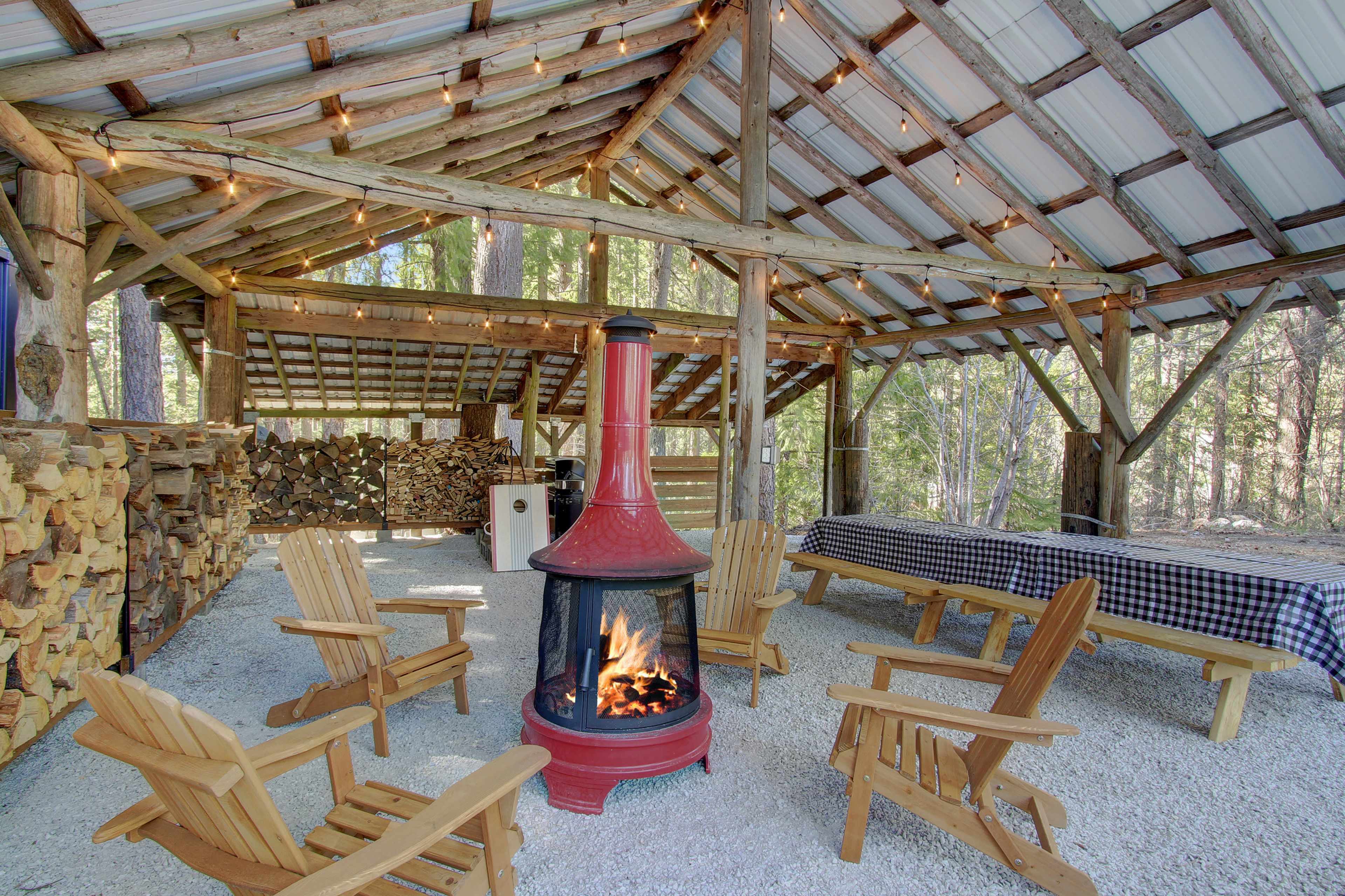 The image shows a rustic outdoor gathering area with wooden chairs surrounding a red metal fireplace, string lights hanging overhead, and a stack of firewood in the background.