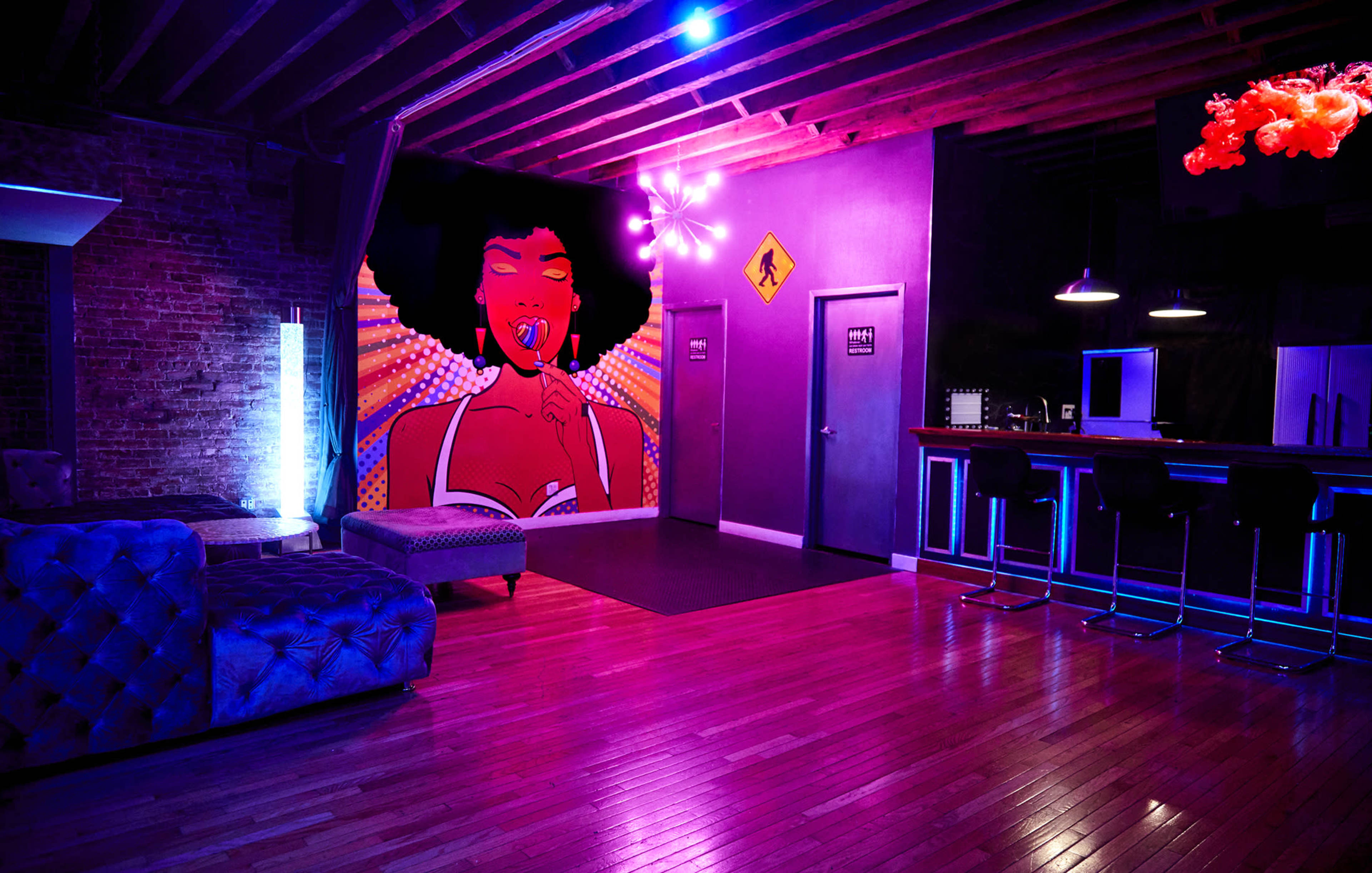 A stylish lounge area featuring a large colorful mural of a woman with an afro on the wall, illuminated by blue and purple lighting, with modern furniture and a bar area.