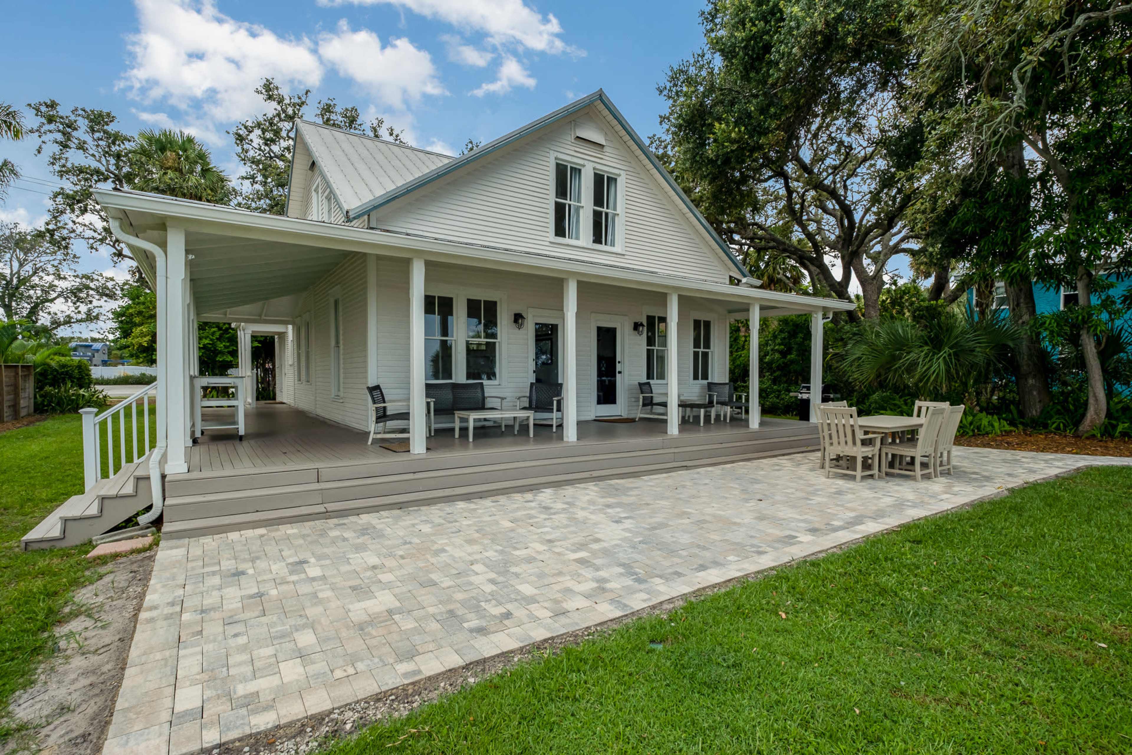 A white two-story house with a porch and paver patio is surrounded by green grass and trees.